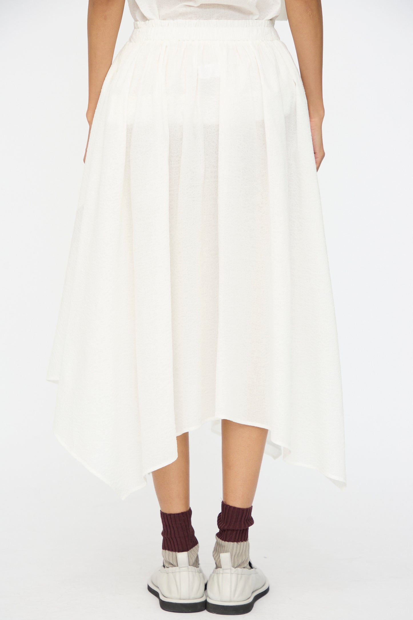 A person is shown from the back against a plain background, wearing the Black Crane Textured Cotton Paper Skirt in White, paired with burgundy socks and white sneakers.