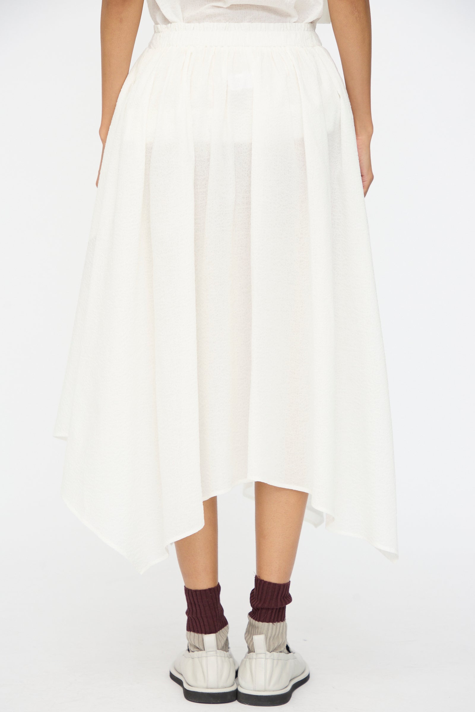 A person is shown from the back against a plain background, wearing the Black Crane Textured Cotton Paper Skirt in White, paired with burgundy socks and white sneakers.