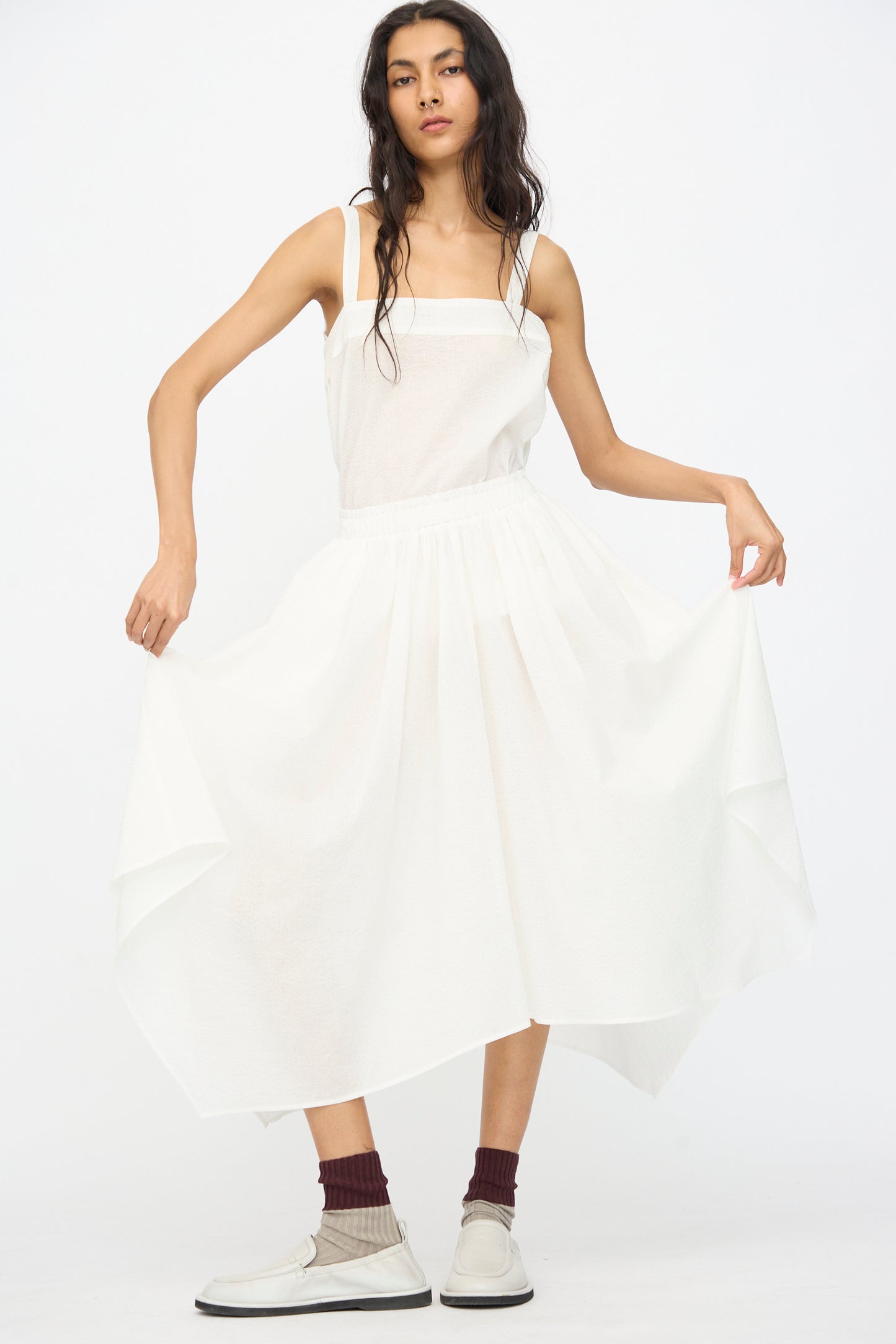 A woman wears the Black Crane Textured Cotton Paper Skirt in White, styled with maroon socks and white shoes as she stands against a plain background, holding out the skirt’s asymmetrical hem.