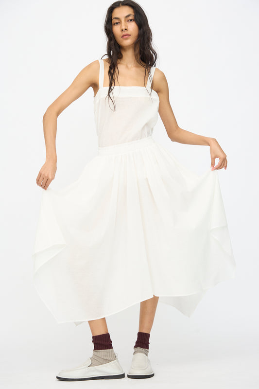 A woman wears the Black Crane Textured Cotton Paper Skirt in White, styled with maroon socks and white shoes as she stands against a plain background, holding out the skirt’s asymmetrical hem.