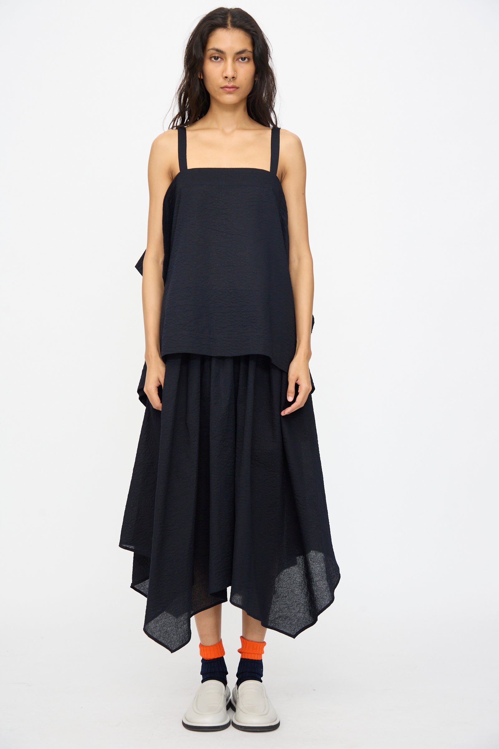 A woman faces forward wearing the Black Crane Textured Cotton Paper Top in Midnight, a loose-fit black tank with square neckline, styled with a matching black midi skirt, orange socks, and white slip-on shoes against a plain white background.