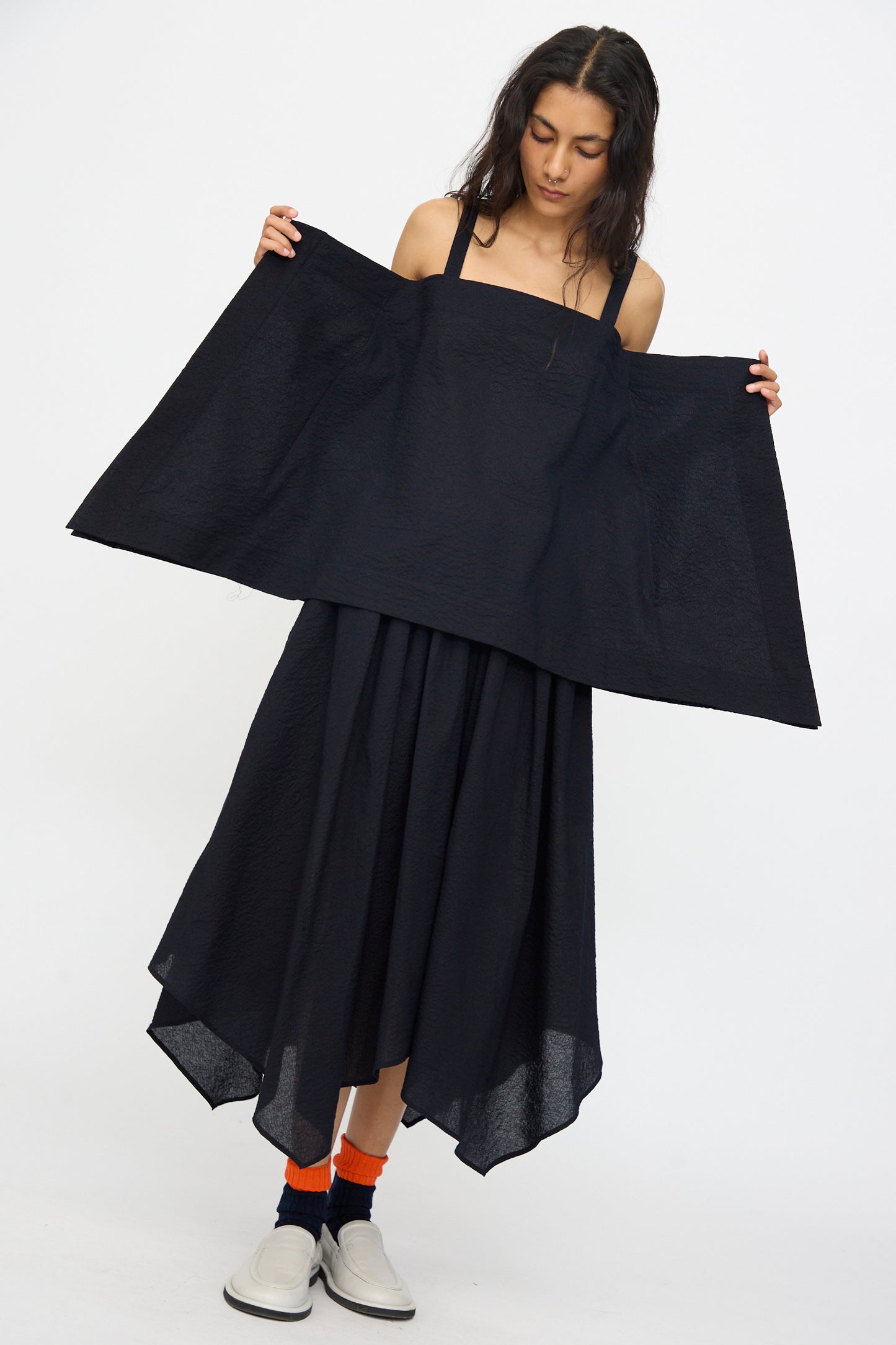 A woman models the Black Crane Textured Cotton Paper Top in Midnight, a loose, asymmetrical Japanese cotton piece. She stands against a plain white background, wearing orange socks and white shoes while holding out part of the garment.