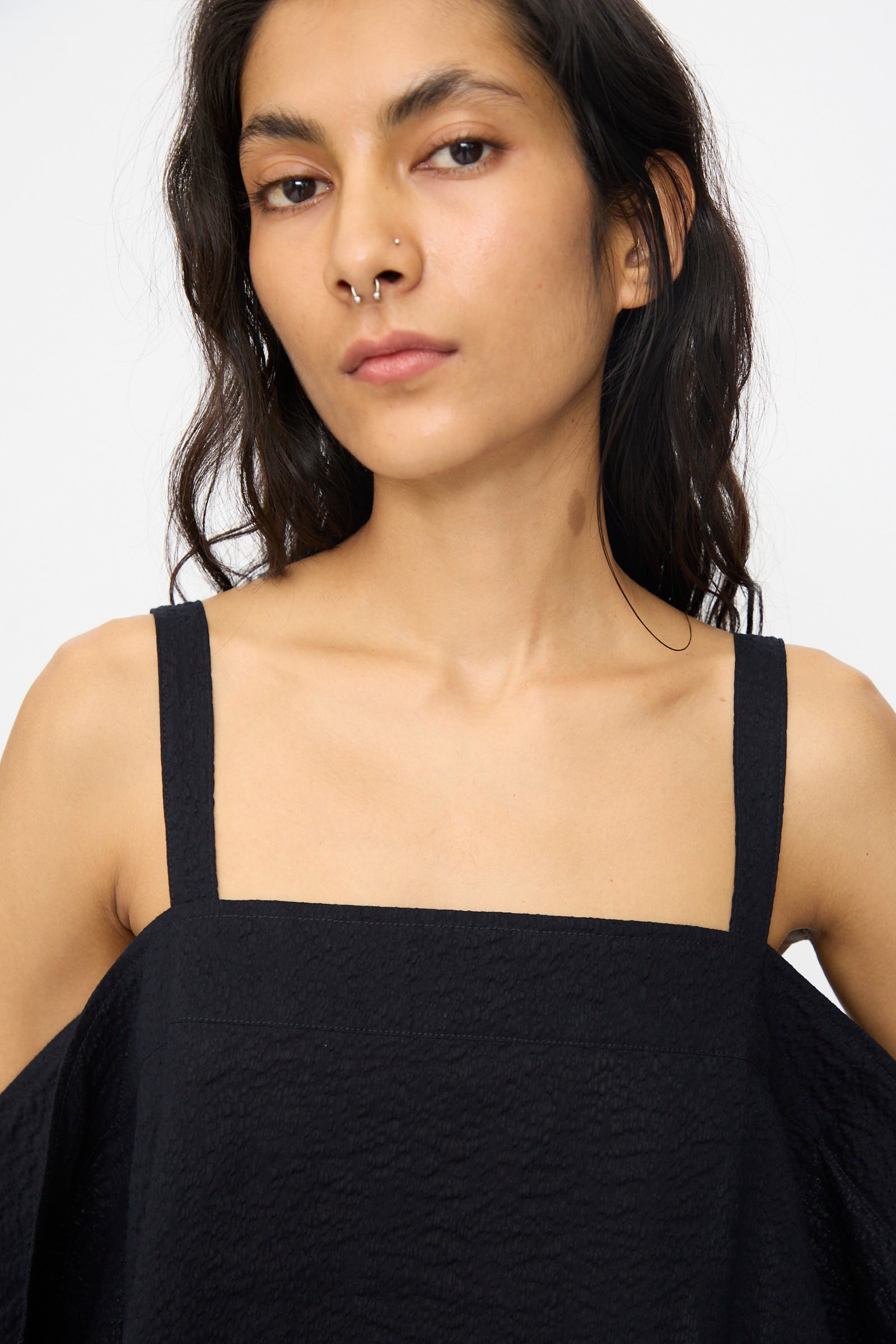 A person with long dark hair and a septum piercing wears the Black Crane Textured Cotton Paper Top in Midnight, featuring a black square neckline, while looking at the camera against a plain background.