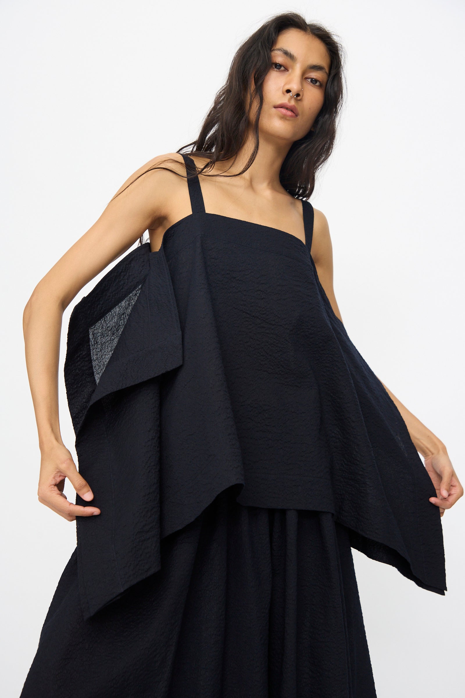 A model wears the Black Crane Textured Cotton Paper Top in Midnight—loose-fitting with wide straps and angled layers—styled with a matching black skirt, against a plain white background.