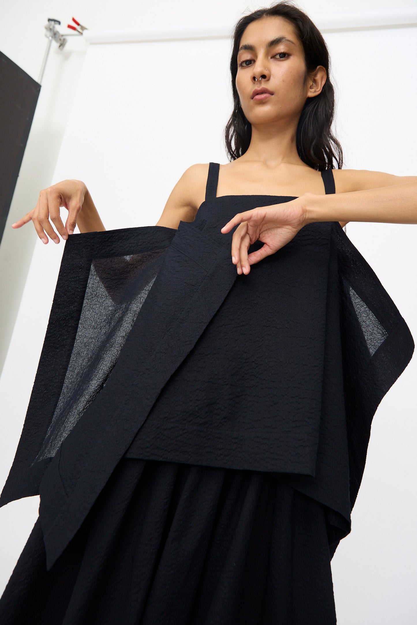 A woman poses against a white background, lifting the structured, layered fabric of the Black Crane Textured Cotton Paper Top in Midnight, which features a square neckline and Japanese-inspired design.