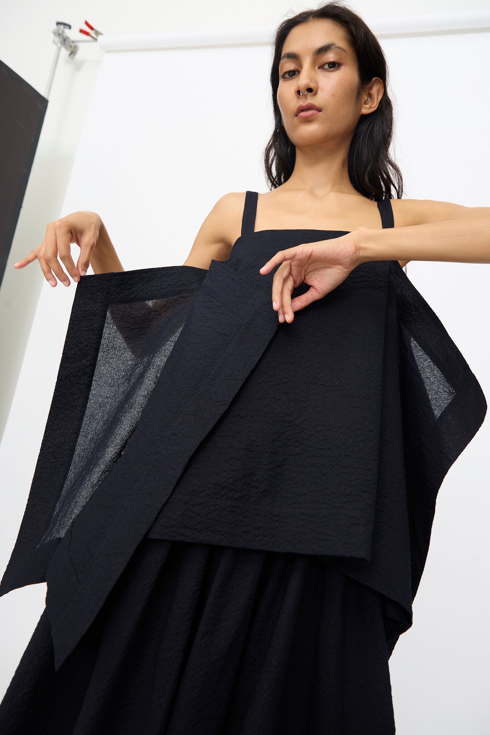 A woman poses against a white background, lifting the structured, layered fabric of the Black Crane Textured Cotton Paper Top in Midnight, which features a square neckline and Japanese-inspired design.