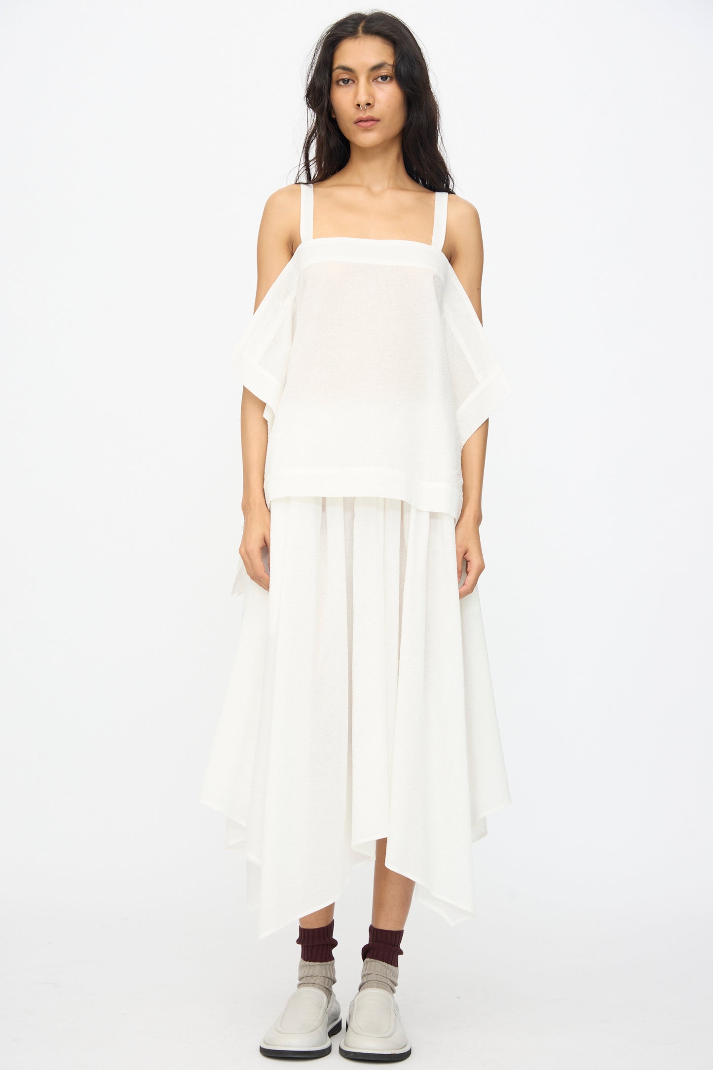 A woman faces forward against a plain white background, wearing the Black Crane Textured Cotton Paper Top in White, a draped white skirt, brown socks, and white slip-on shoes.