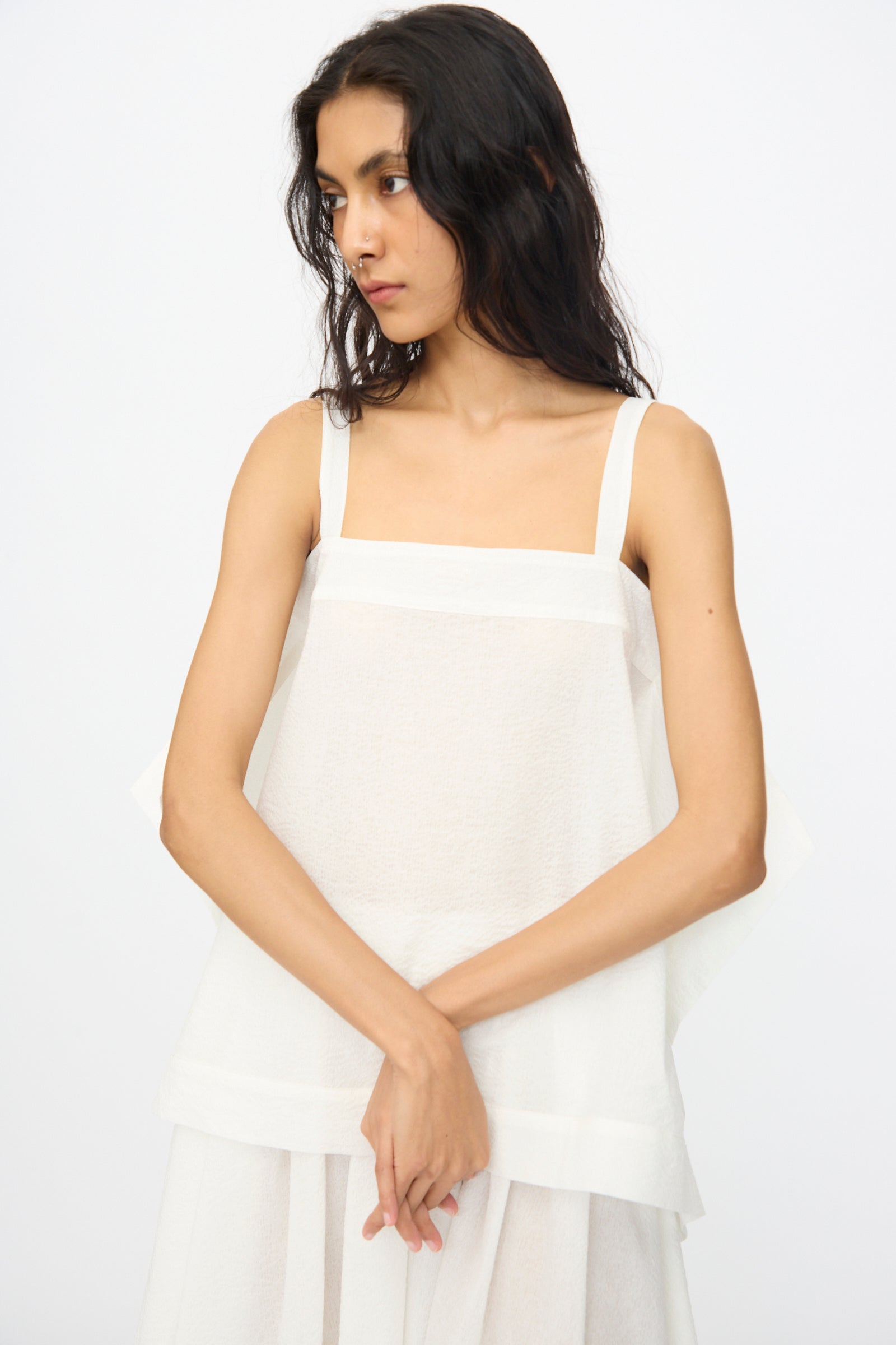 A woman with long dark hair wears the Black Crane Textured Cotton Paper Top in White, a sleeveless and loose-fitting piece, standing against a plain light background and looking slightly to her left with hands clasped.