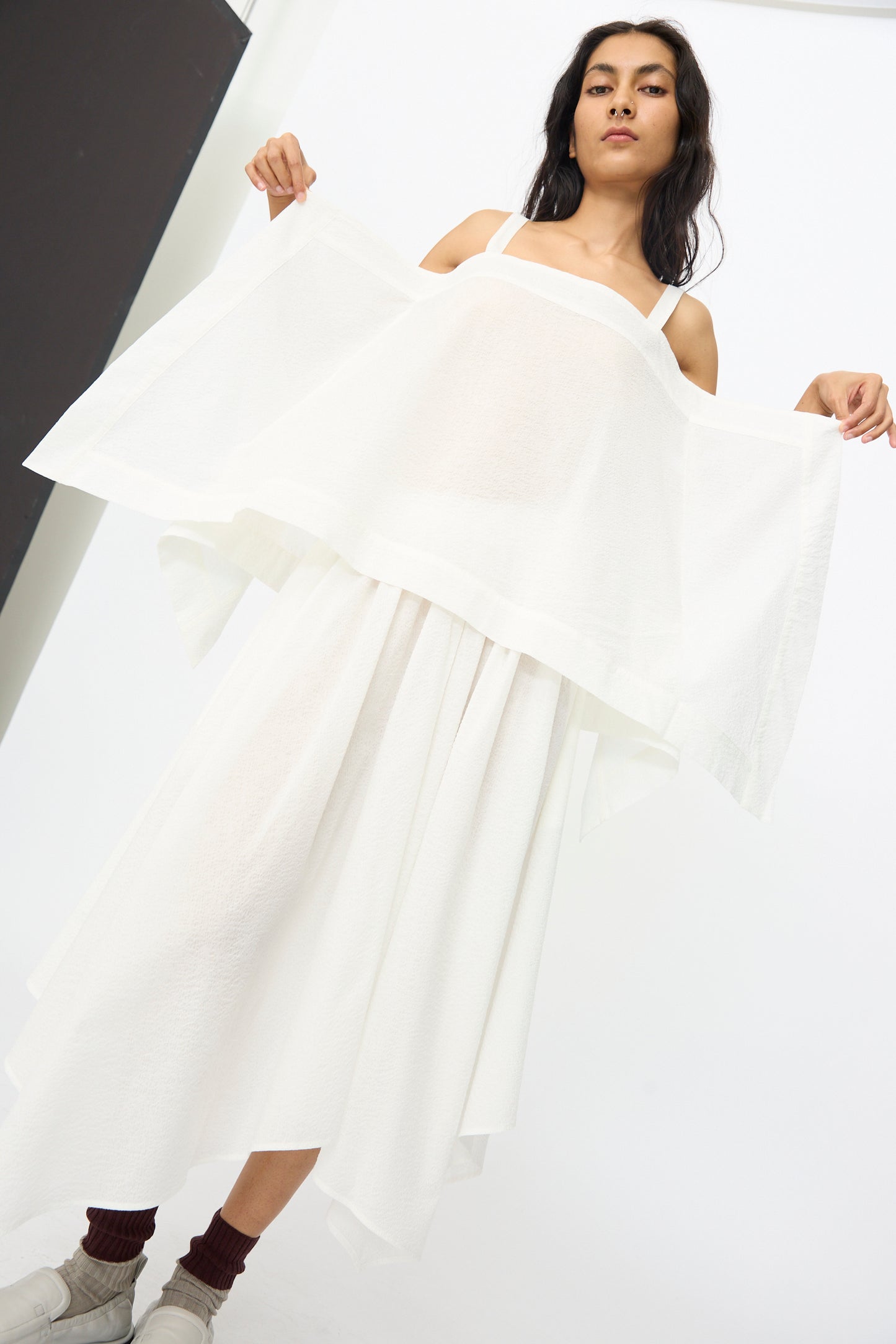 A person stands indoors wearing Black Crane’s Textured Cotton Paper Top in White, styled with white sneakers and maroon socks, holding out the sides of the lightweight, flowy top.