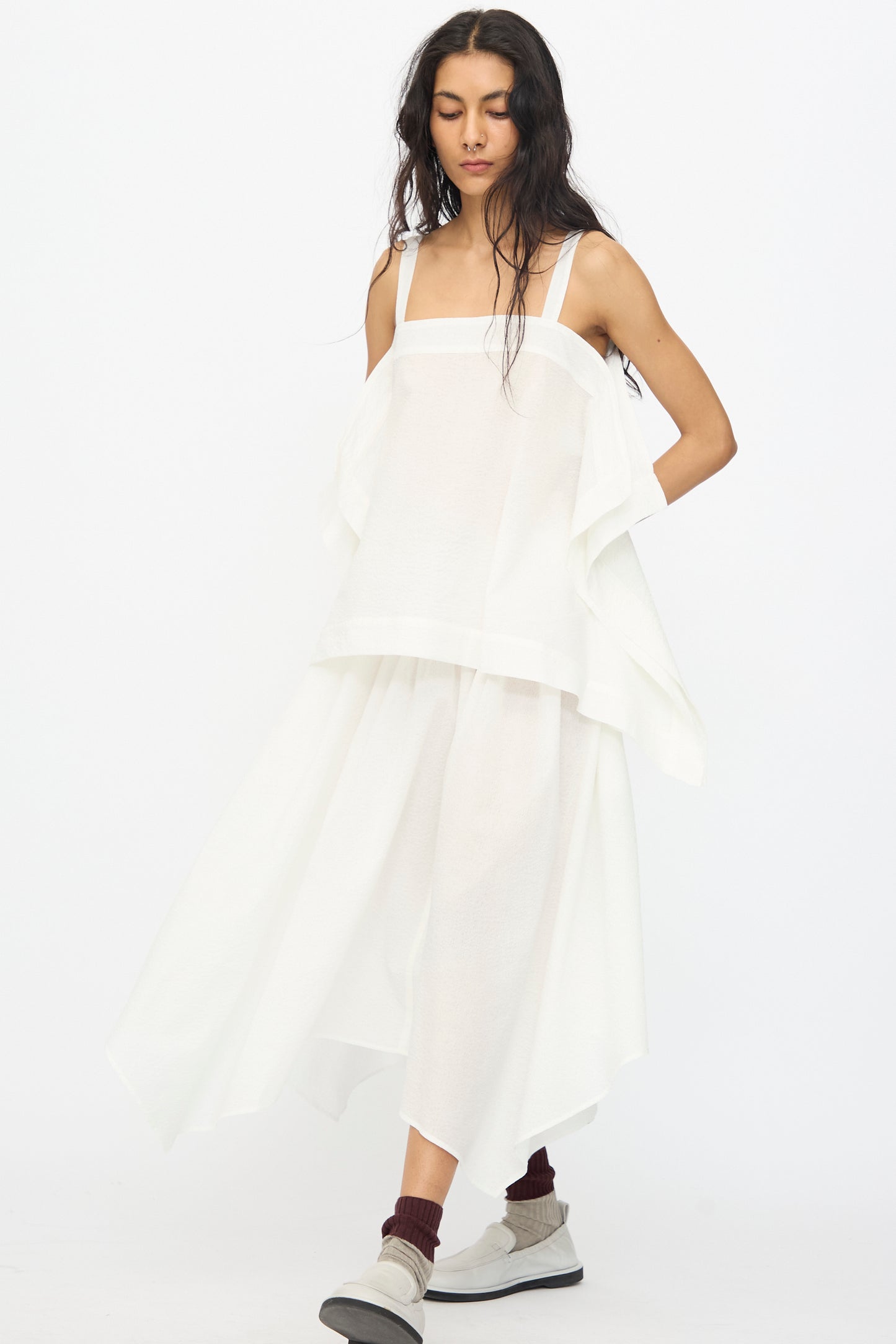 A woman wears the Black Crane Textured Cotton Paper Top in White, styled with a matching asymmetrical skirt, maroon socks, and white sneakers, standing against a plain white background.