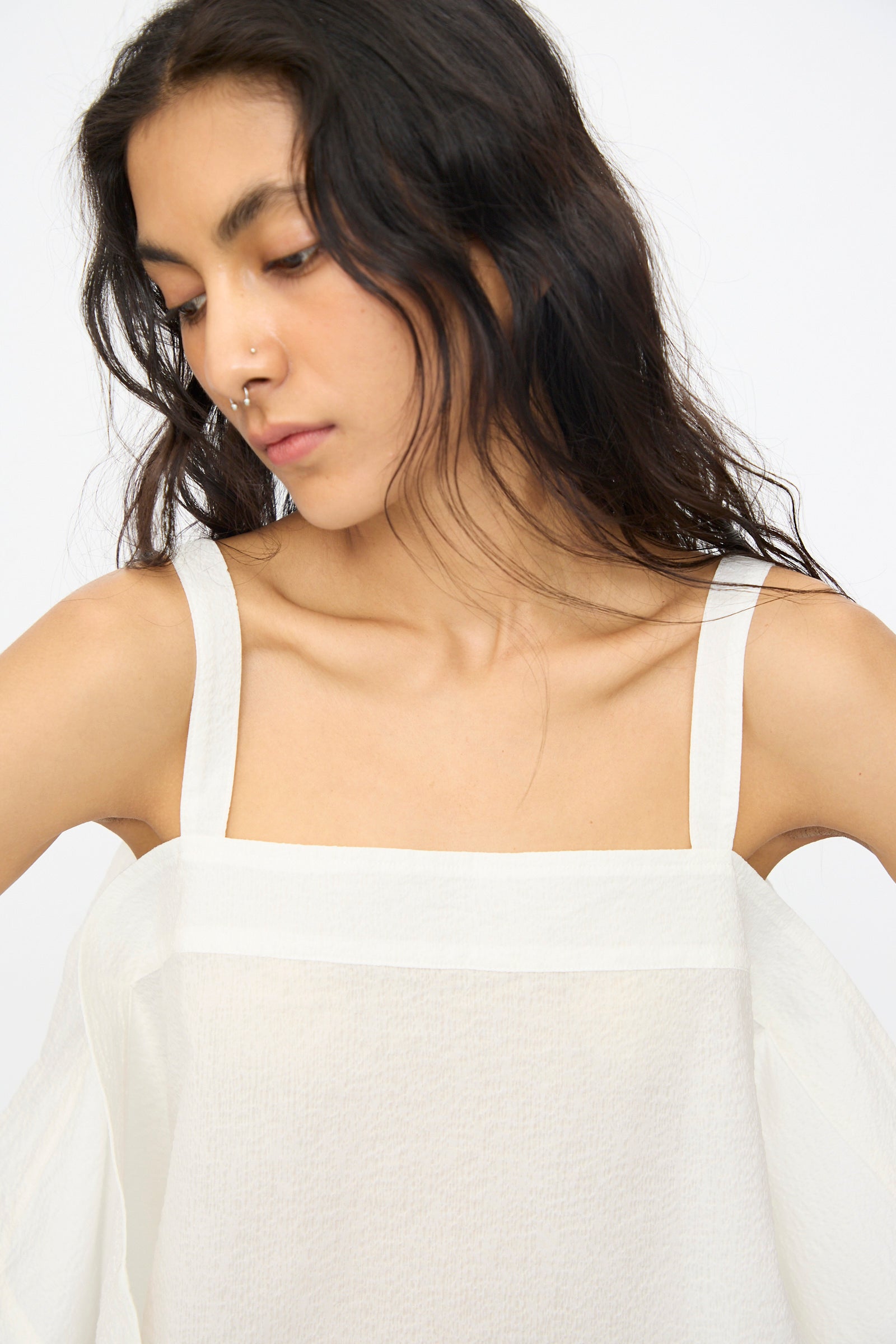 A person with long dark hair and a nose ring wears the Black Crane Textured Cotton Paper Top in White, featuring a square neckline, while looking down against a plain background.