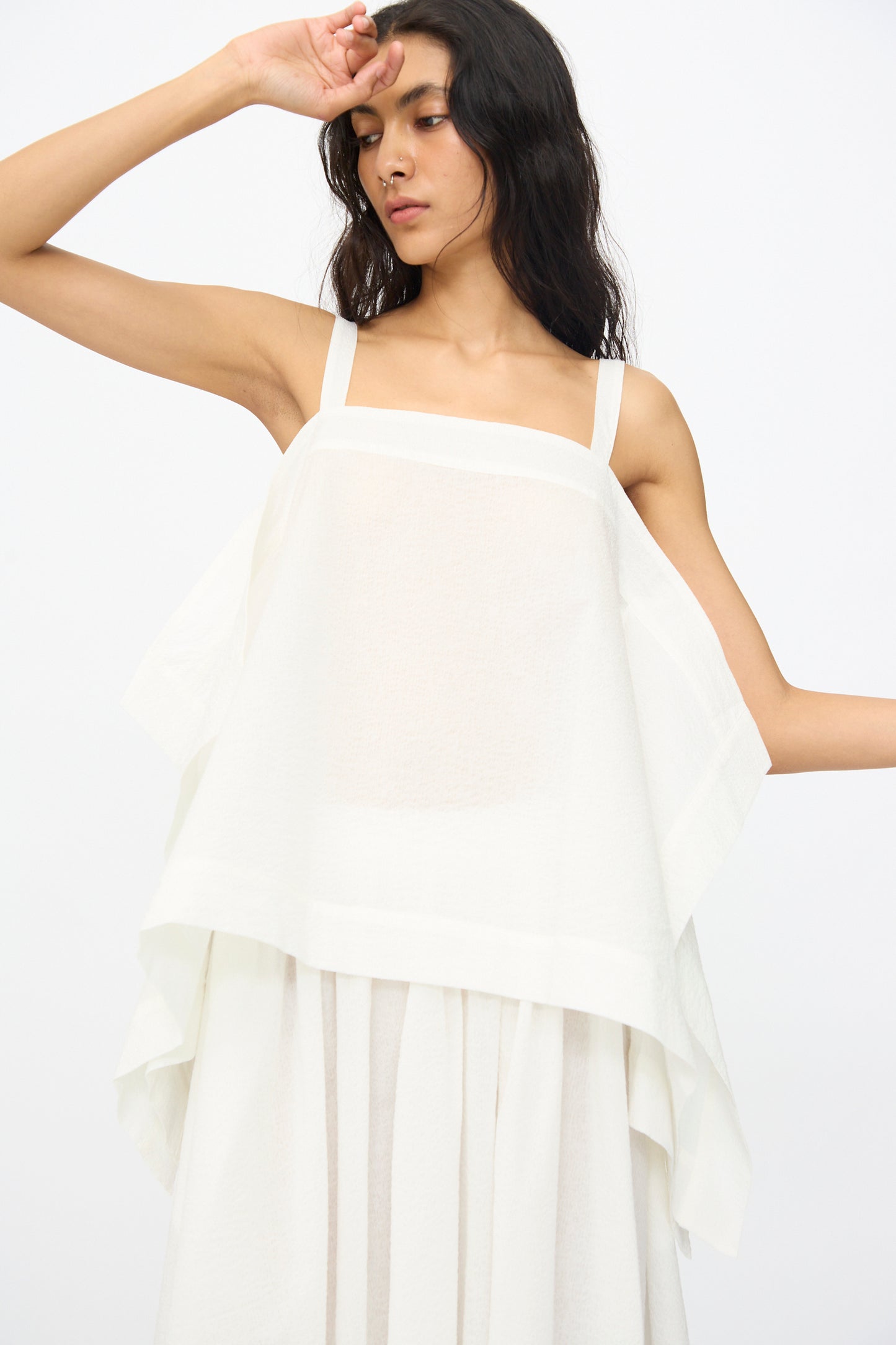 A woman in the Black Crane Textured Cotton Paper Top in White—a sleeveless, lightweight design made from Japanese textured cotton—stands against a plain white background with one arm raised to her forehead.