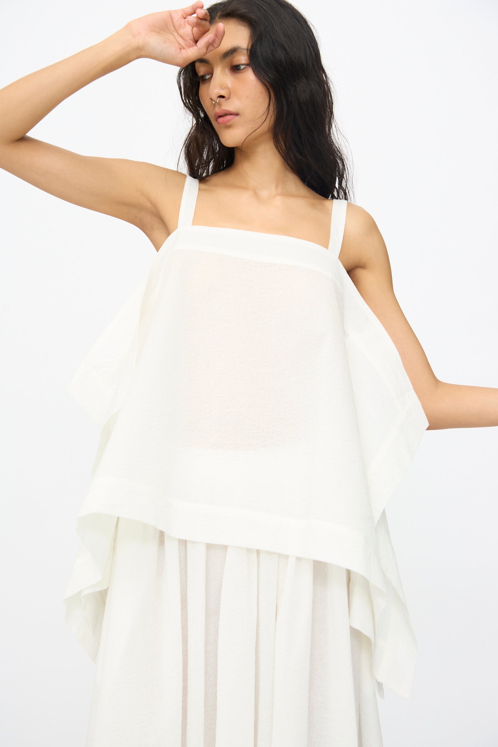 A woman in the Black Crane Textured Cotton Paper Top in White—a sleeveless, lightweight design made from Japanese textured cotton—stands against a plain white background with one arm raised to her forehead.
