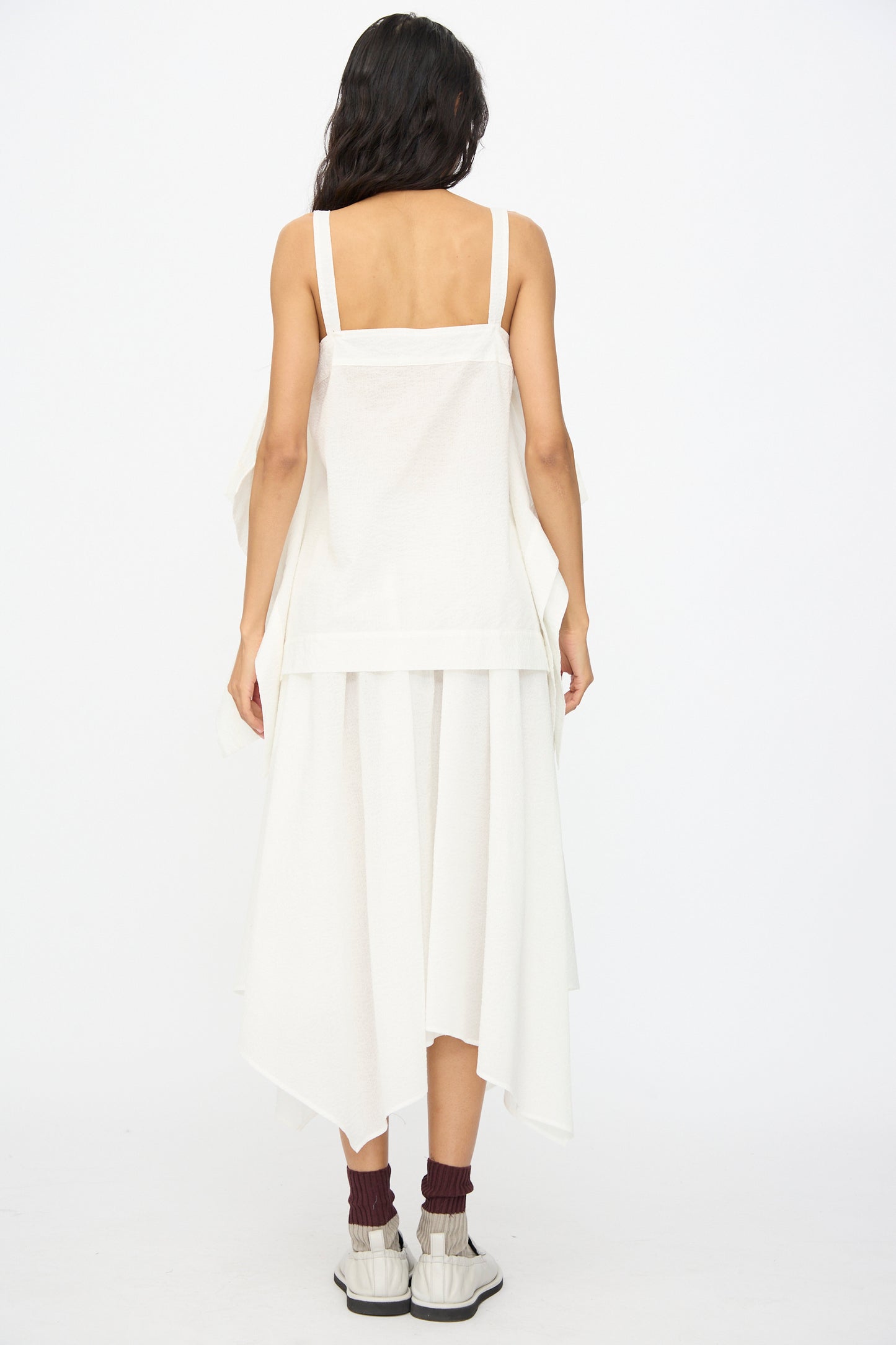 A woman with long dark hair models the Black Crane Textured Cotton Paper Top in White, styled with a white asymmetrical skirt, maroon socks, and white sandals, photographed from the back against a plain background.