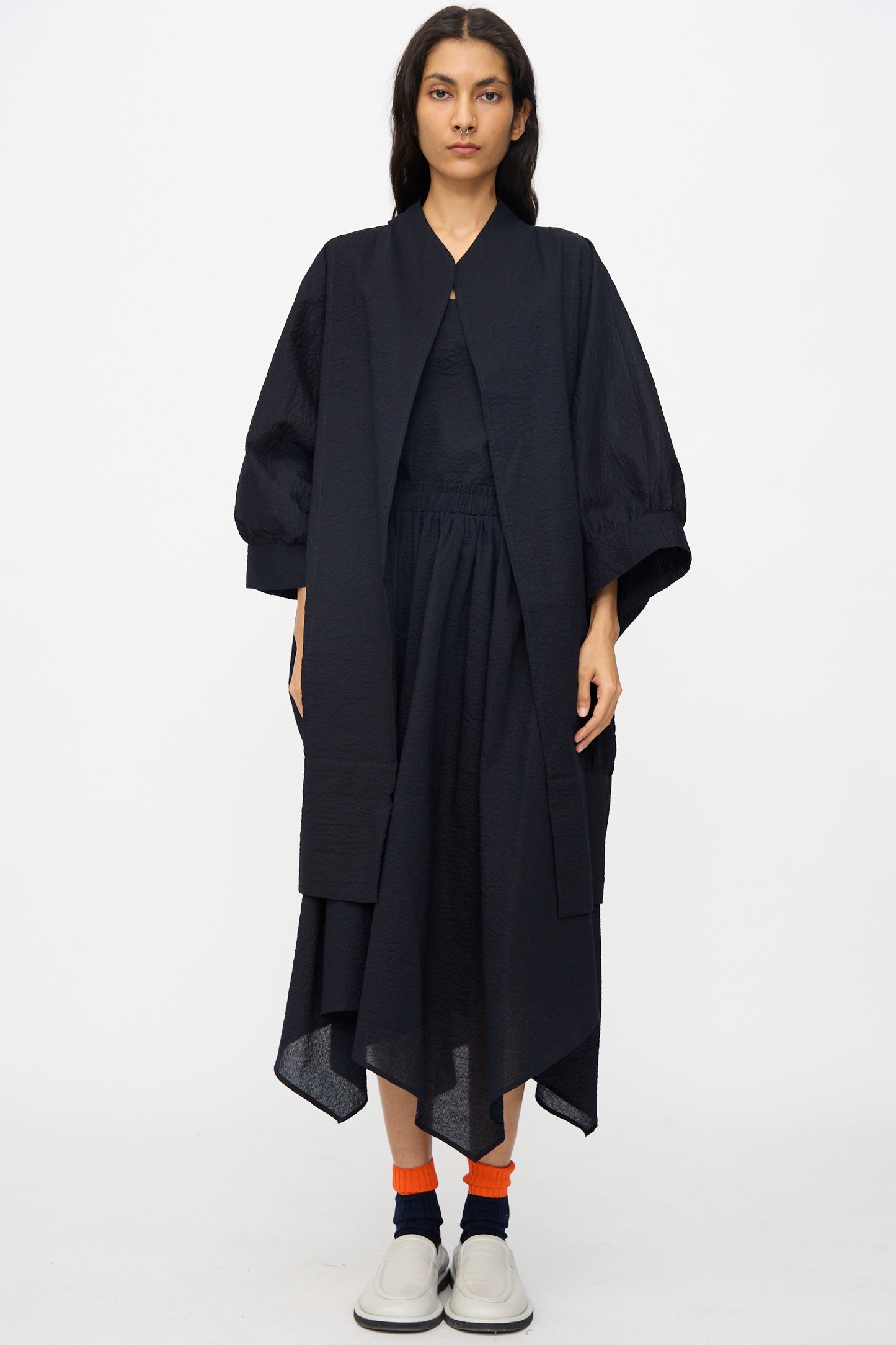 A person faces forward against a plain white background, wearing Black Crane’s Textured Cotton Spoon Jacket in Midnight styled with a black skirt, orange socks, and white shoes for an oversized look.