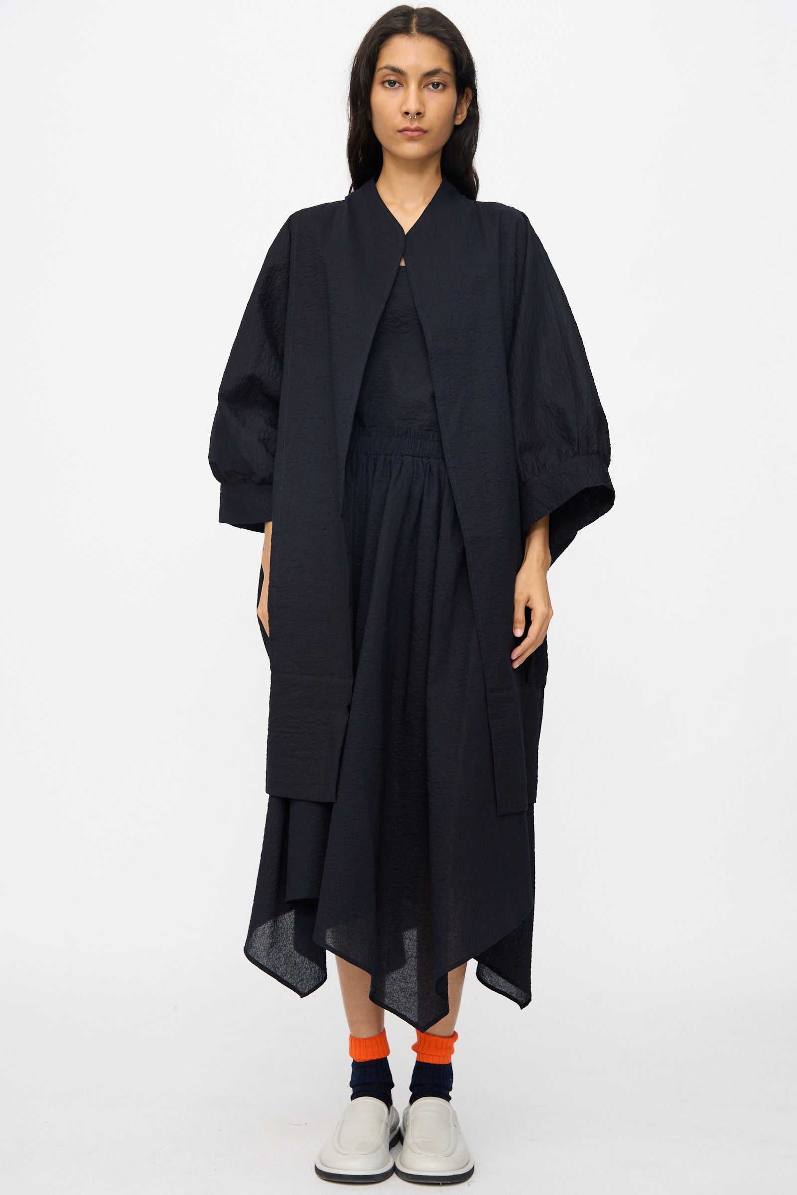 A person faces forward against a plain white background, wearing Black Crane’s Textured Cotton Spoon Jacket in Midnight styled with a black skirt, orange socks, and white shoes for an oversized look.