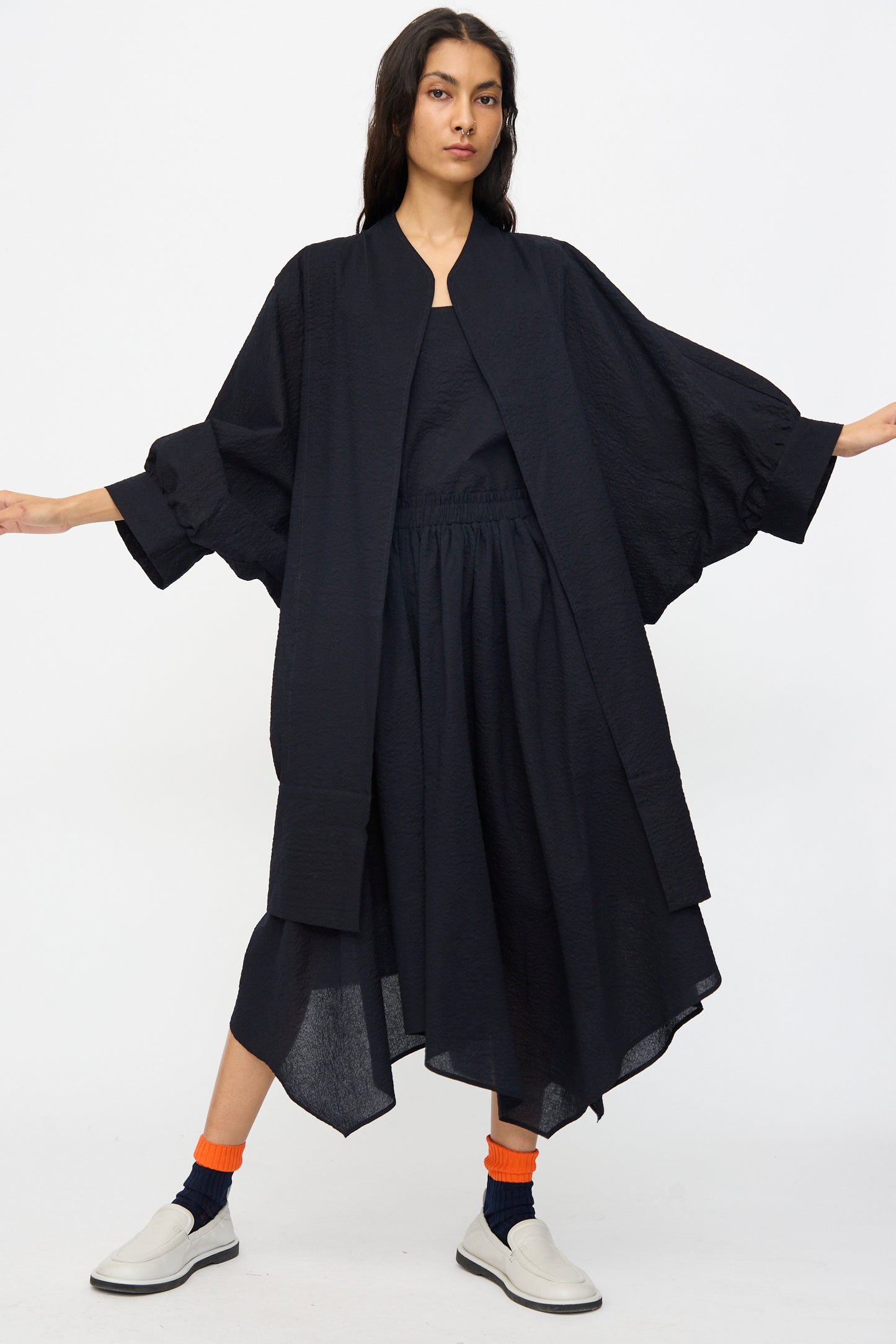 A person stands with arms outstretched, wearing the Black Crane Textured Cotton Spoon Jacket in Midnight, styled loosely like a kimono, paired with a matching skirt, orange socks, and white slip-on shoes against a plain white background.