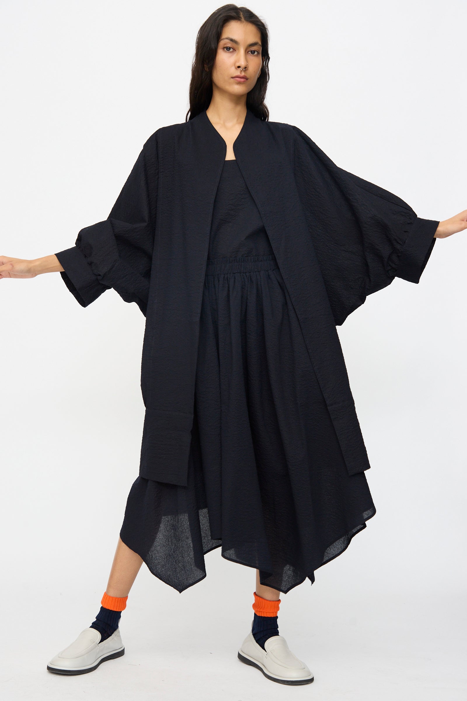 A person stands with arms outstretched, wearing the Black Crane Textured Cotton Spoon Jacket in Midnight, styled loosely like a kimono, paired with a matching skirt, orange socks, and white slip-on shoes against a plain white background.