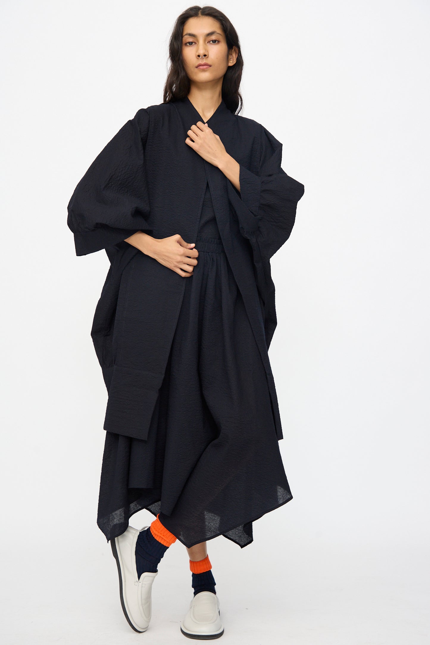 A person stands against a plain background wearing the Black Crane Textured Cotton Spoon Jacket in Midnight, paired with orange and navy socks and white slip-on shoes.