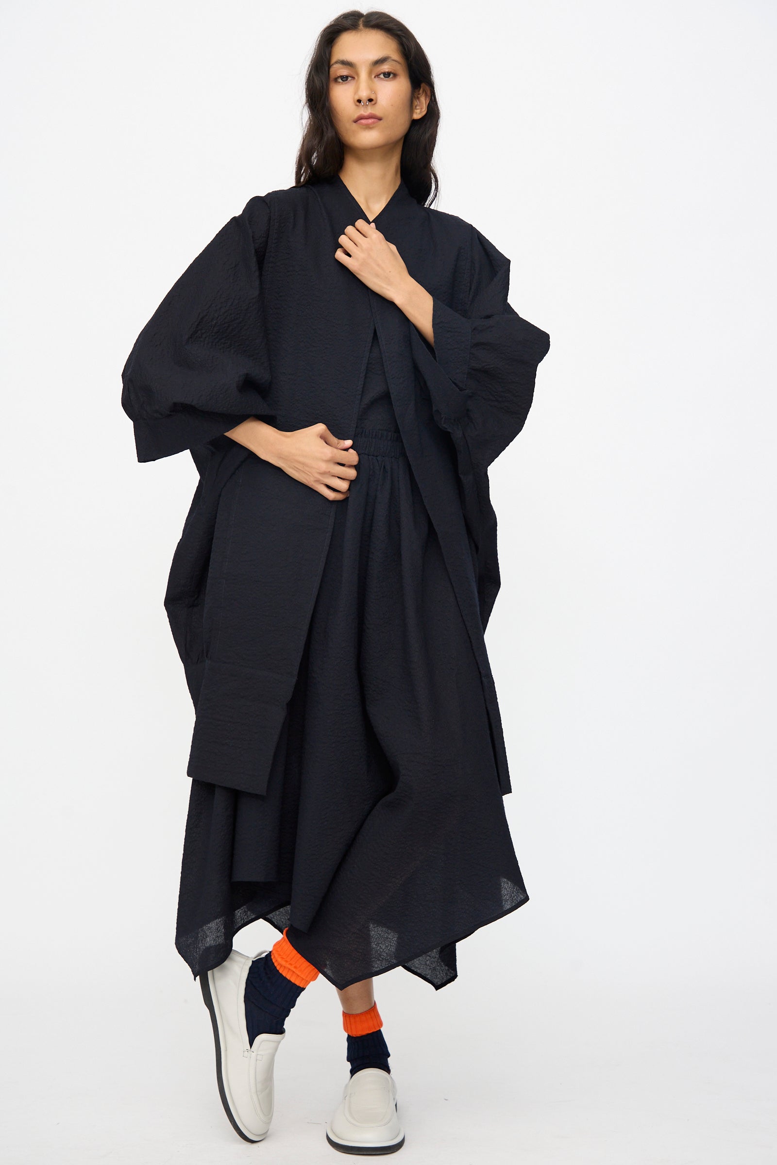 A person stands against a plain background wearing the Black Crane Textured Cotton Spoon Jacket in Midnight, paired with orange and navy socks and white slip-on shoes.