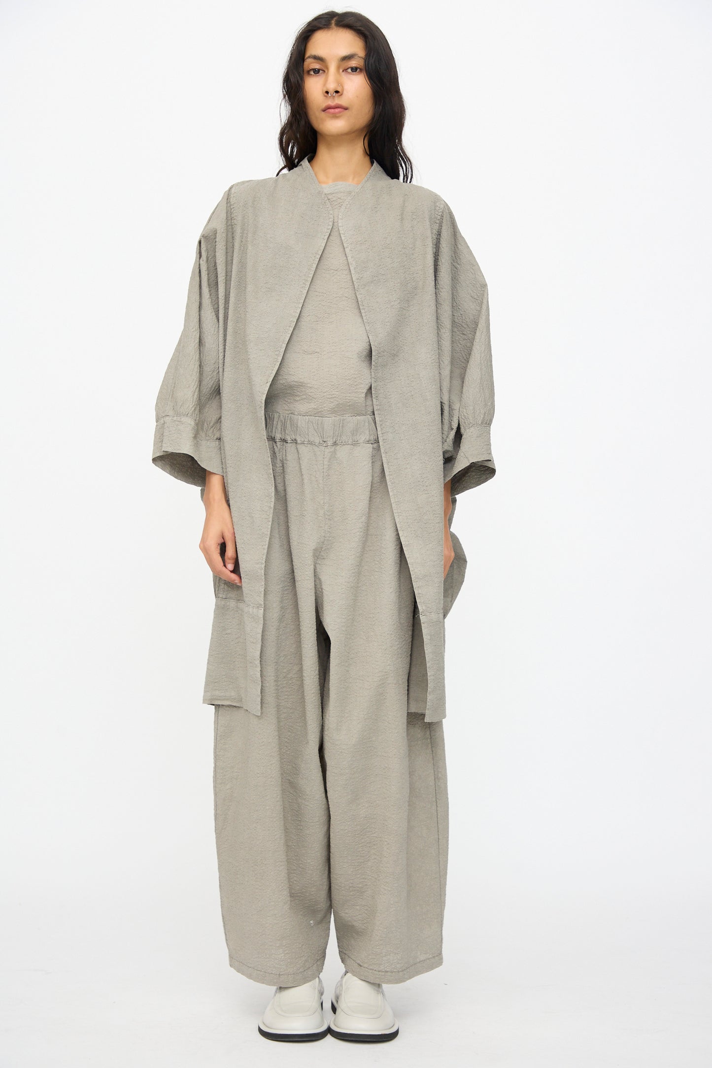 A person stands against a white background wearing the Black Crane Textured Cotton Spoon Jacket in Pale Grey with wide-leg pants, a matching top, and white shoes.