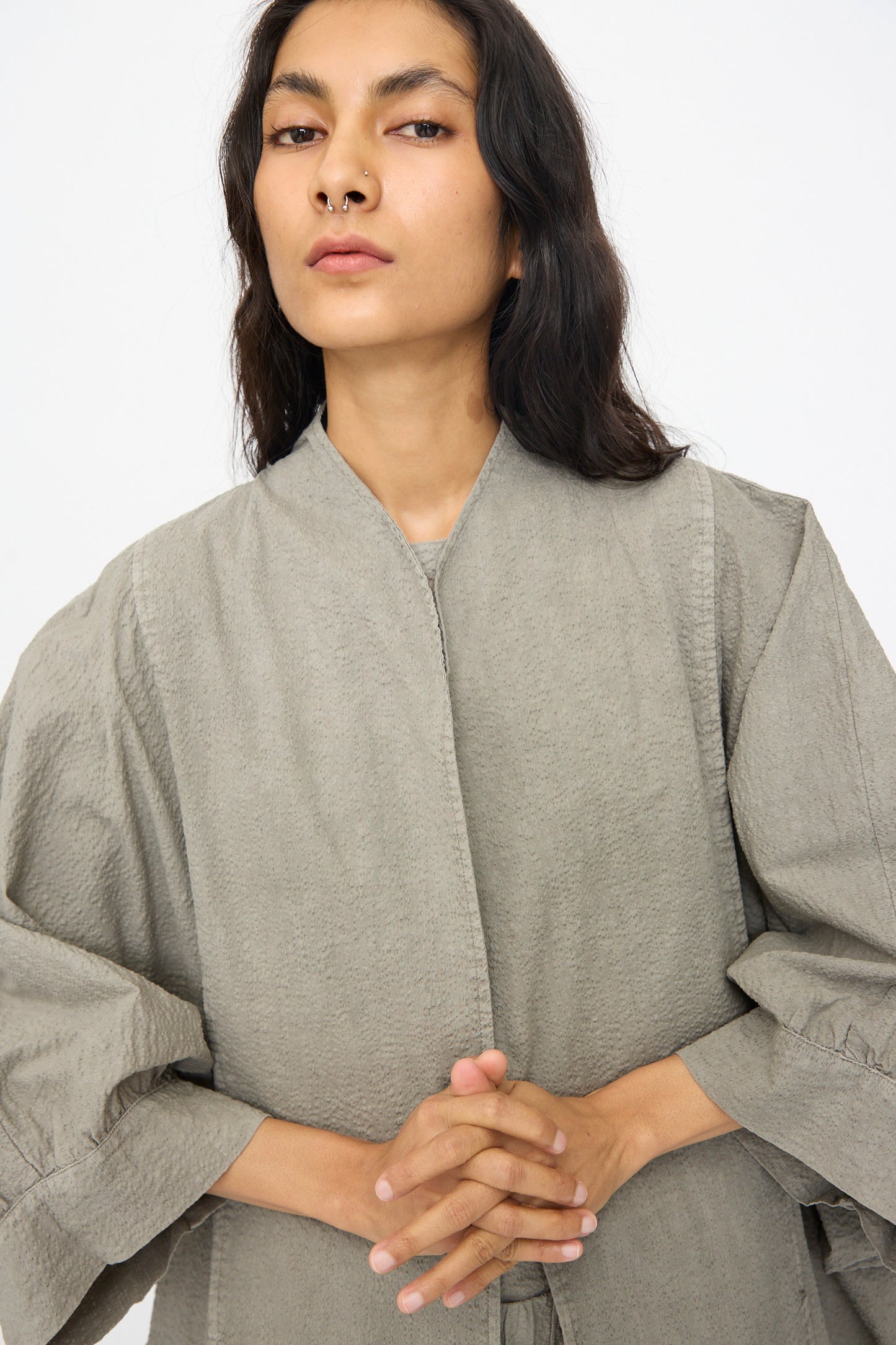A person with long dark hair and a nose ring wears the Black Crane Textured Cotton Spoon Jacket in Pale Grey, standing with hands clasped and looking at the camera against a plain background.
