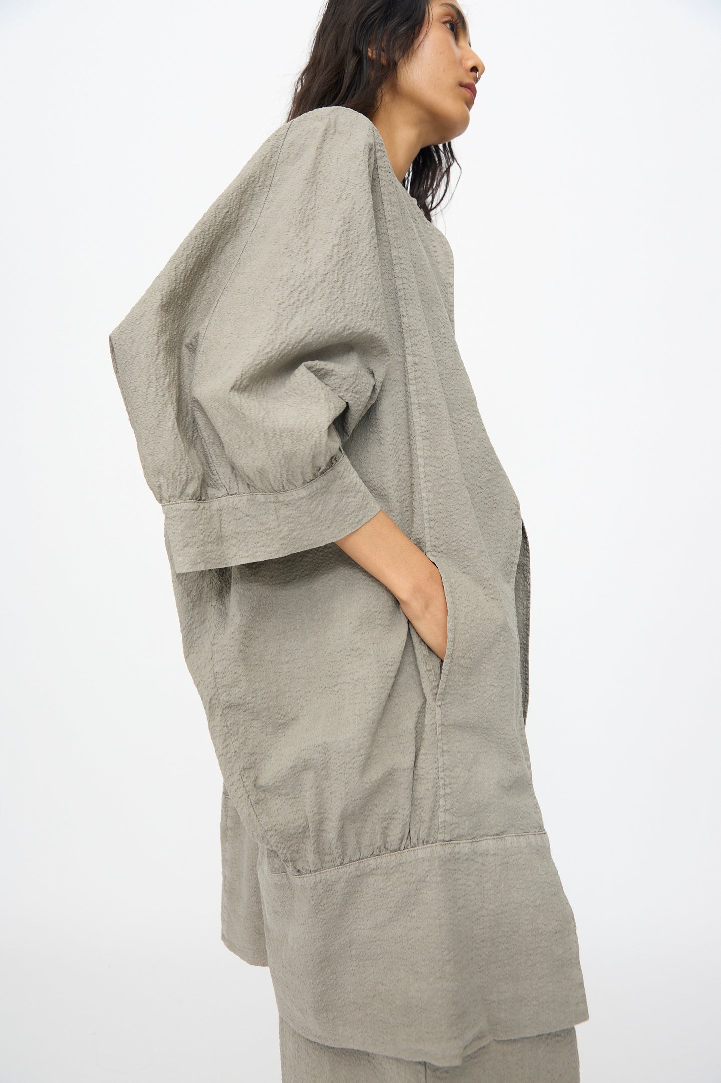 A person wears the Black Crane Textured Cotton Spoon Jacket in Pale Grey—an oversized, lightweight Japanese textured cotton jacket with wide sleeves and pockets—while standing against a plain white background.