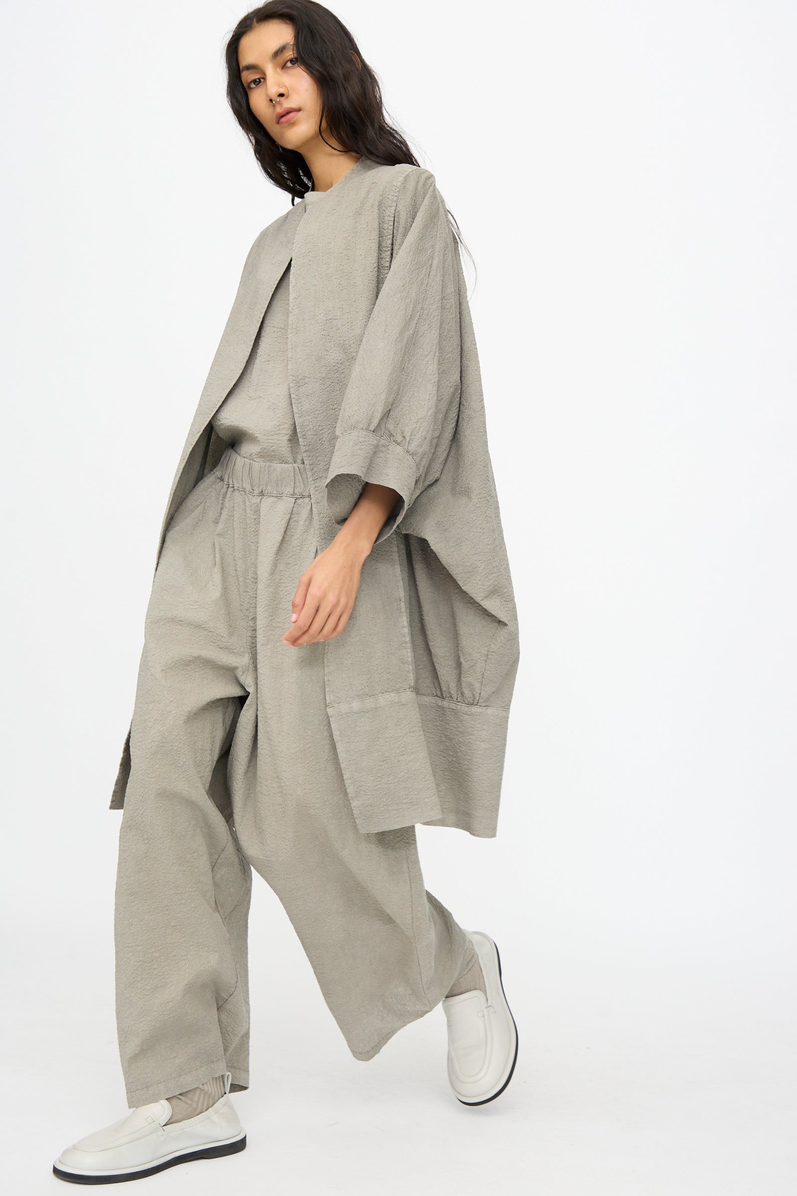 A person wears the Black Crane Textured Cotton Spoon Jacket in Pale Grey, styled with wide-leg pants and white slip-on shoes, walking against a plain white background.