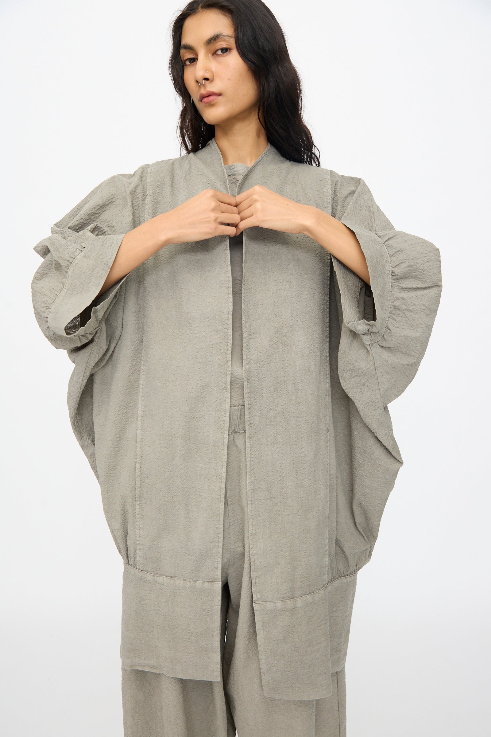 A person stands against a plain background, wearing the Black Crane Textured Cotton Spoon Jacket in Pale Grey—an oversized kimono-style design with wide sleeves—adjusting the front of this Japanese textured cotton piece.