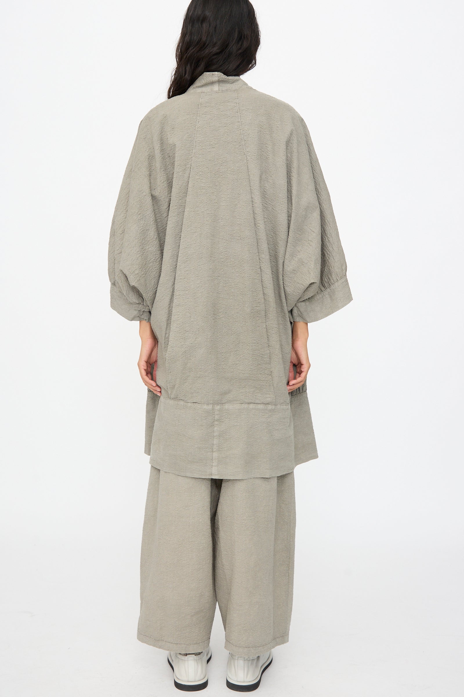 A person stands with their back to the camera, wearing the Black Crane Textured Cotton Spoon Jacket in Pale Grey—an oversized top paired with matching wide-leg pants and white shoes—set against a plain white background.