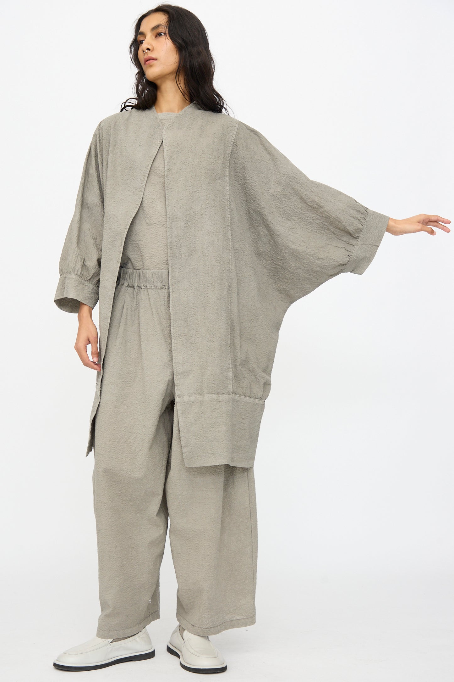 Against a plain background, a person wears Black Crane's Textured Cotton Spoon Jacket in Pale Grey—an oversized fit with long sleeves, paired with wide pants and white shoes for a minimalist look.