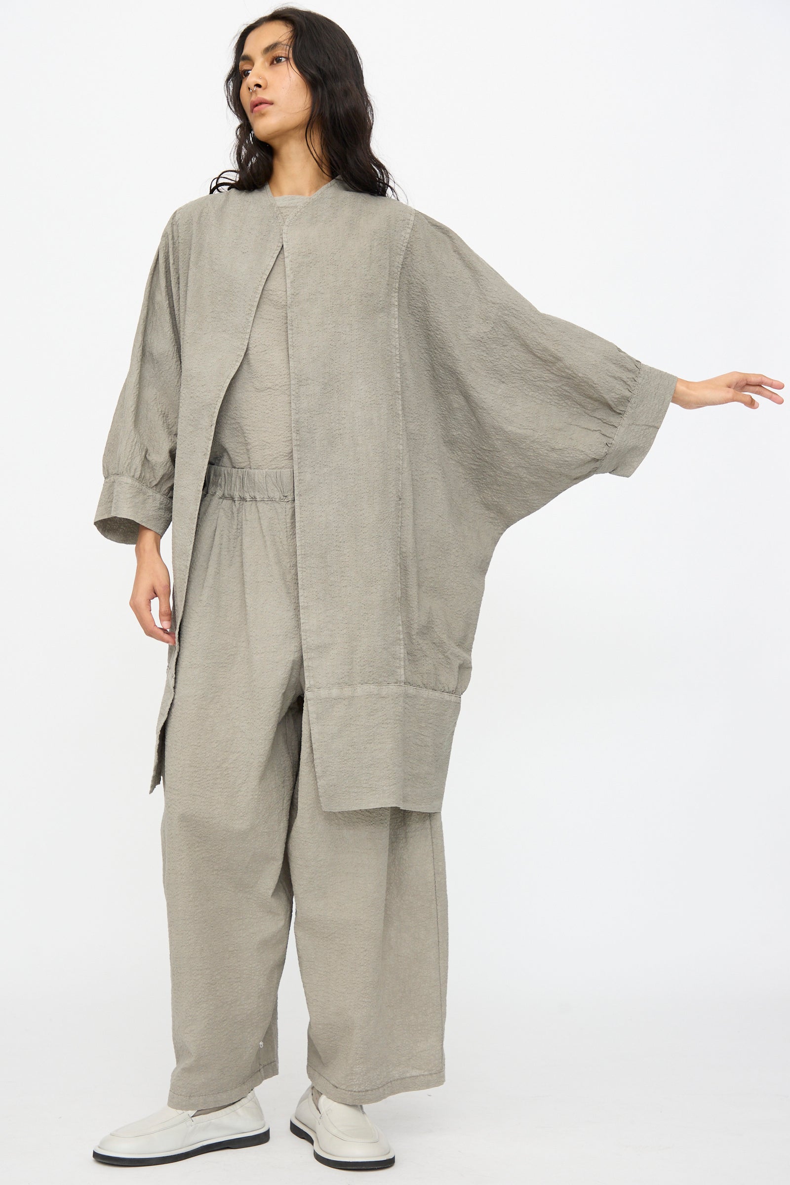 Against a plain background, a person wears Black Crane's Textured Cotton Spoon Jacket in Pale Grey—an oversized fit with long sleeves, paired with wide pants and white shoes for a minimalist look.