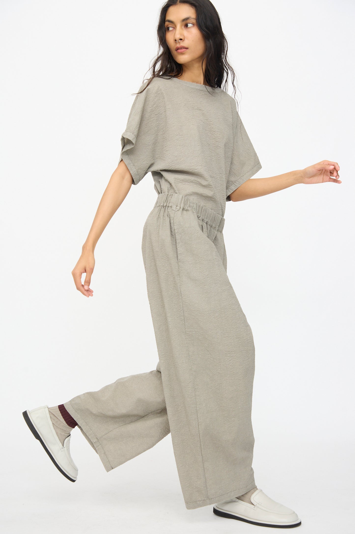 A person walks confidently against a plain white background, dressed in Black Crane’s Textured Cotton Wide Pants in Pale Grey and a loose-fitting, light gray short-sleeve top.