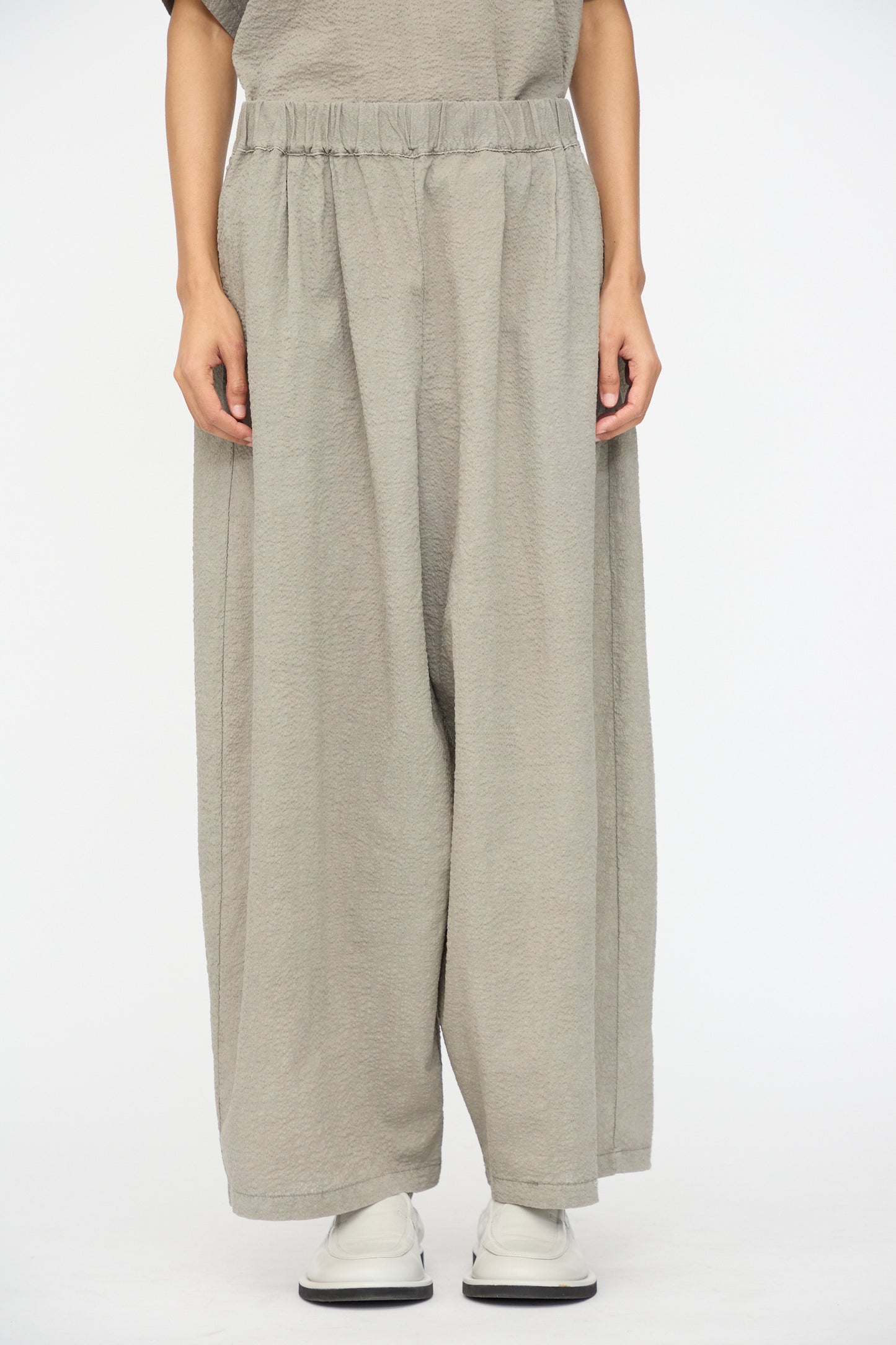 A person wears Black Crane's Textured Cotton Wide Pants in Pale Grey, styled with white shoes and standing against a plain white backdrop.