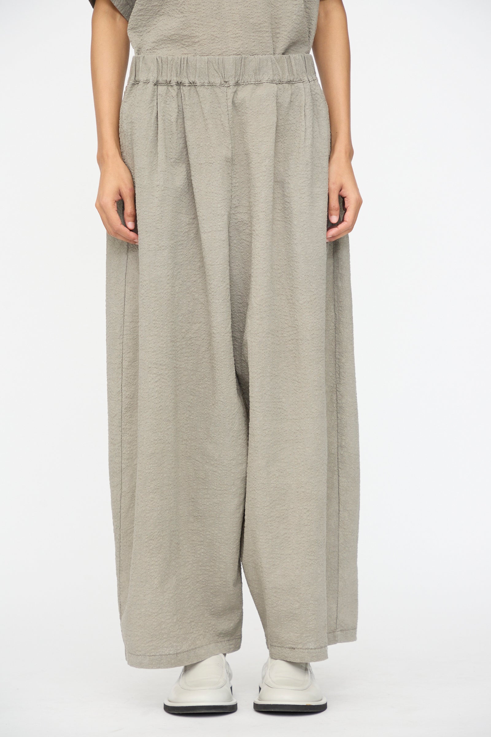 A person wears Black Crane's Textured Cotton Wide Pants in Pale Grey, styled with white shoes and standing against a plain white backdrop.