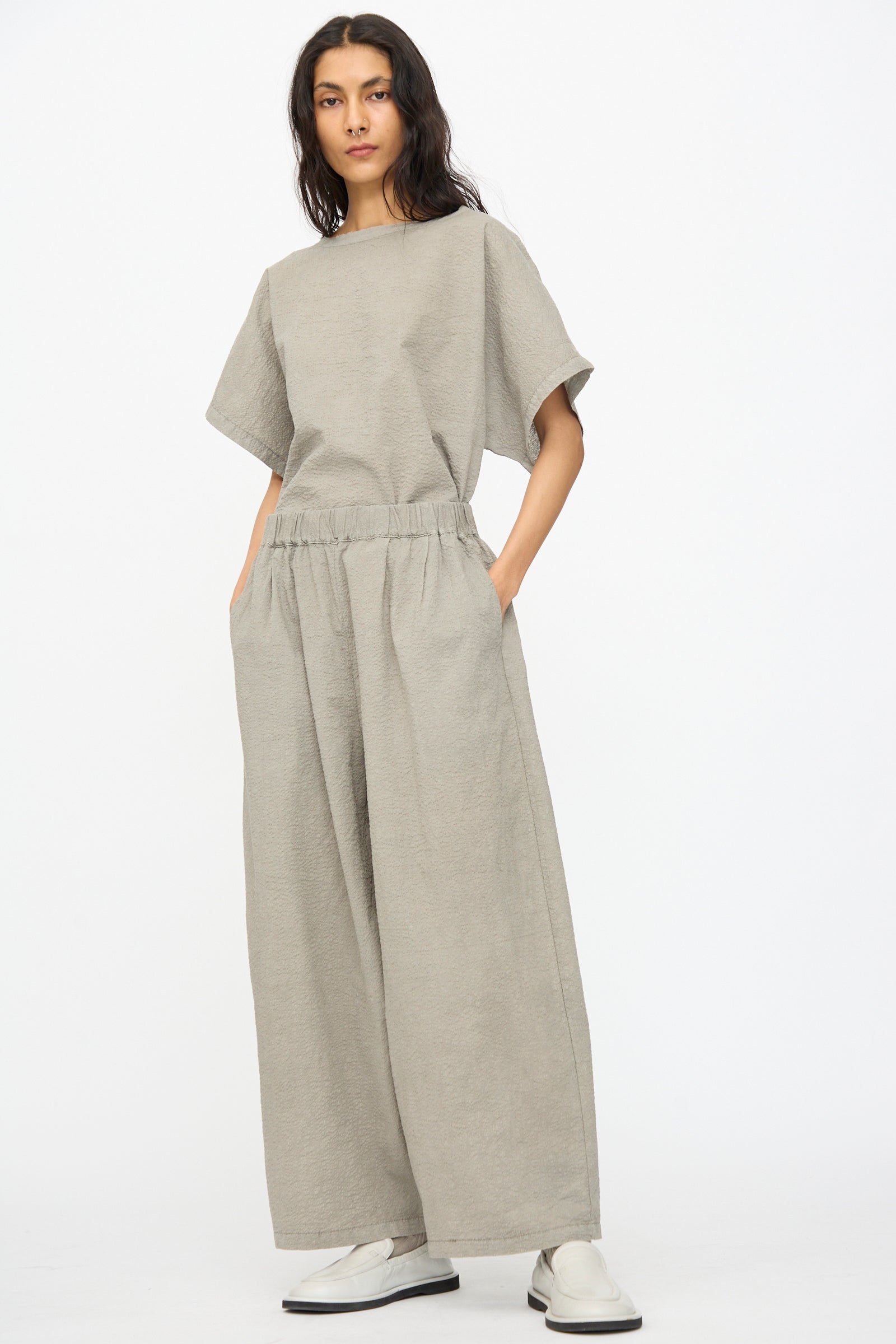 A person stands against a plain background wearing Black Crane’s Textured Cotton Wide Pants in Pale Grey, paired with a loose-fitting light gray short-sleeve top and white slip-on shoes.
