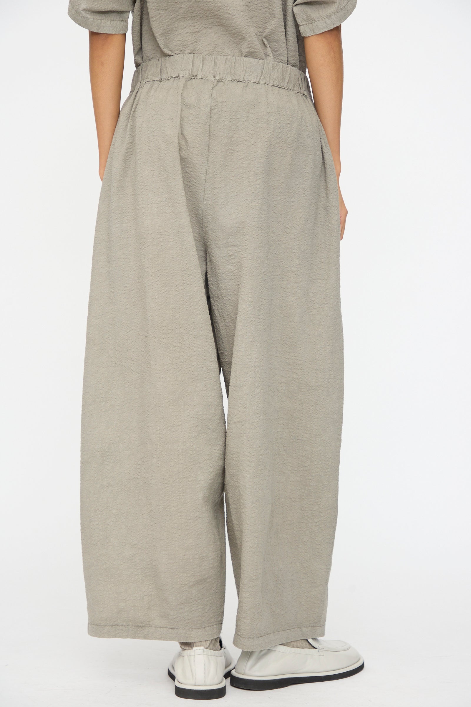 Person wearing Black Crane Textured Cotton Wide Pants in Pale Grey with white sneakers, photographed from behind against a plain background.