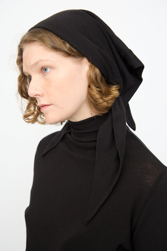 A person with light skin and short curly hair wears the Black Crane Triangle Scarf in Black—a soft cotton cashmere blend—paired with a black turtleneck, looking to the left against a plain white background. The scarf is thoughtfully made in the USA.