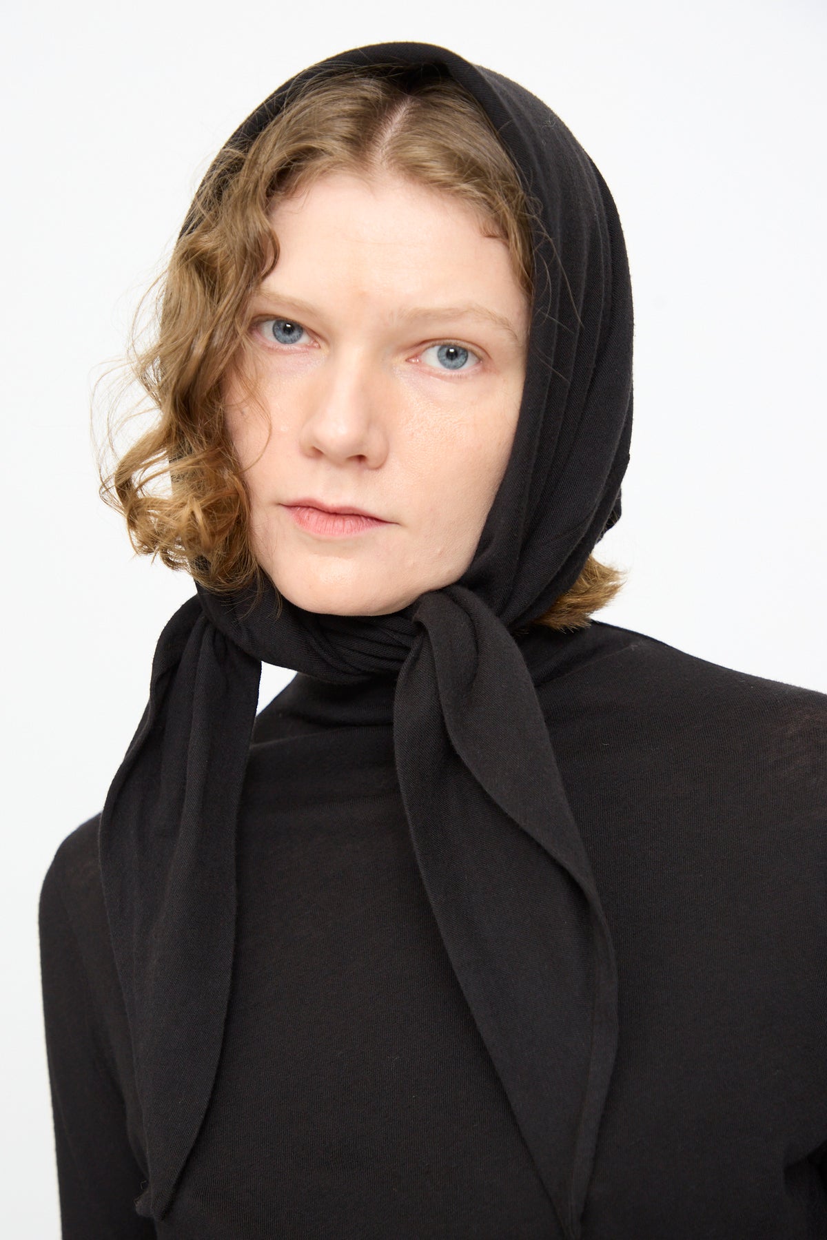 A person with light skin and wavy blonde hair wears the Black Crane Triangle Scarf in Black and a black top, looking directly at the camera against a plain white background.