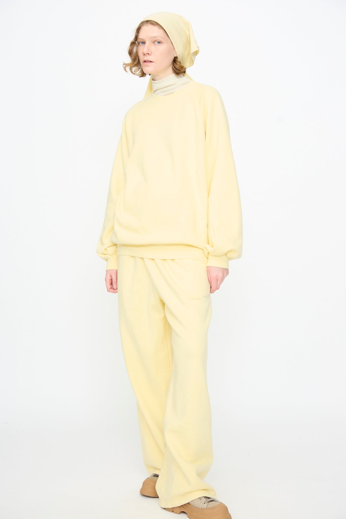 A person stands against a white background, dressed in a loose-fitting, pale yellow outfit paired with the Black Crane Triangle Scarf in Custard and beige shoes. Crafted from a soft cotton cashmere blend, the ensemble exudes both comfort and sophistication.
