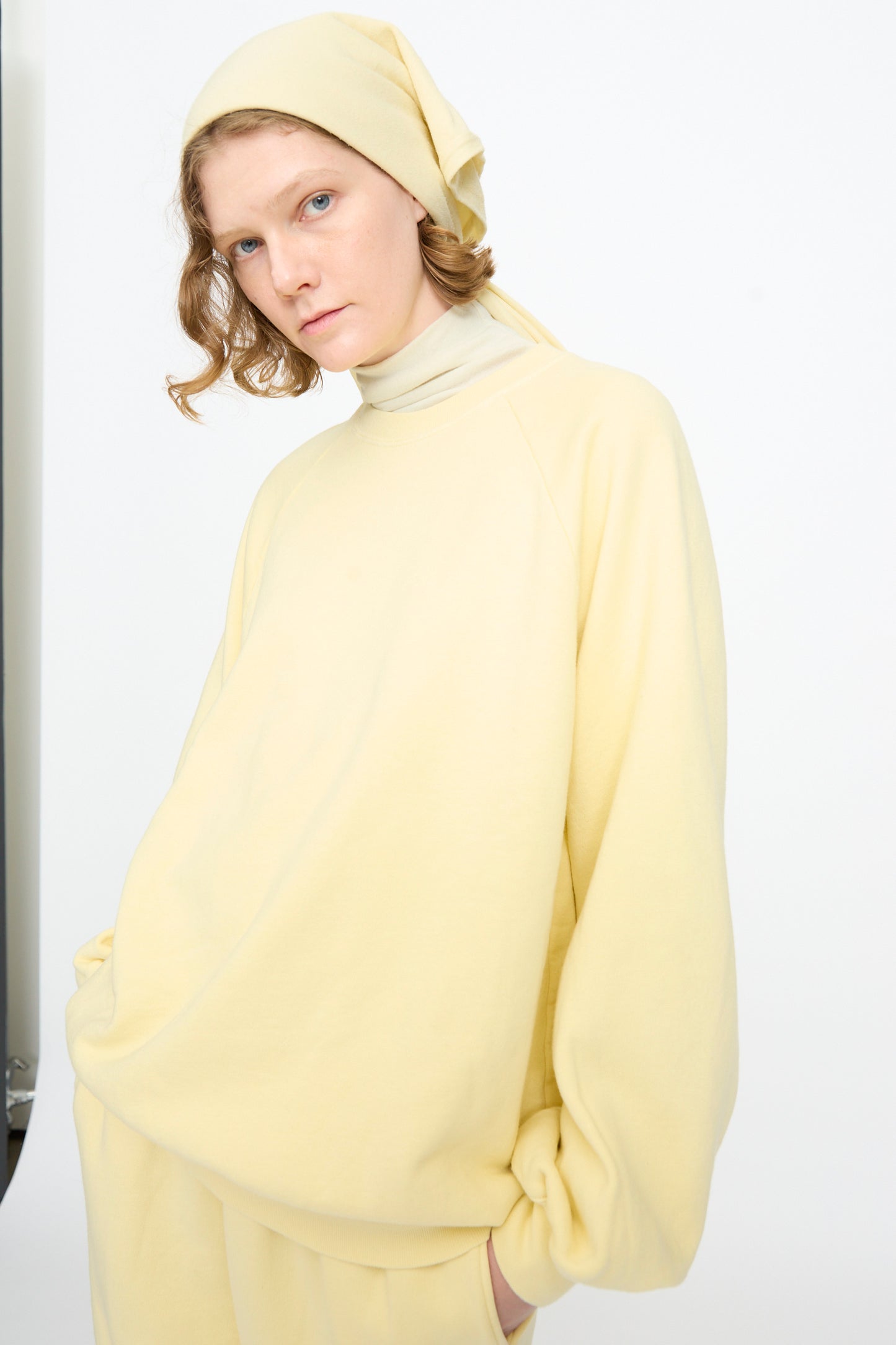 A person stands against a plain white background with one hand in a pocket, wearing a light yellow oversized sweatshirt and matching pants, styled with the Black Crane Triangle Scarf in Custard crafted from a soft cotton-cashmere blend.