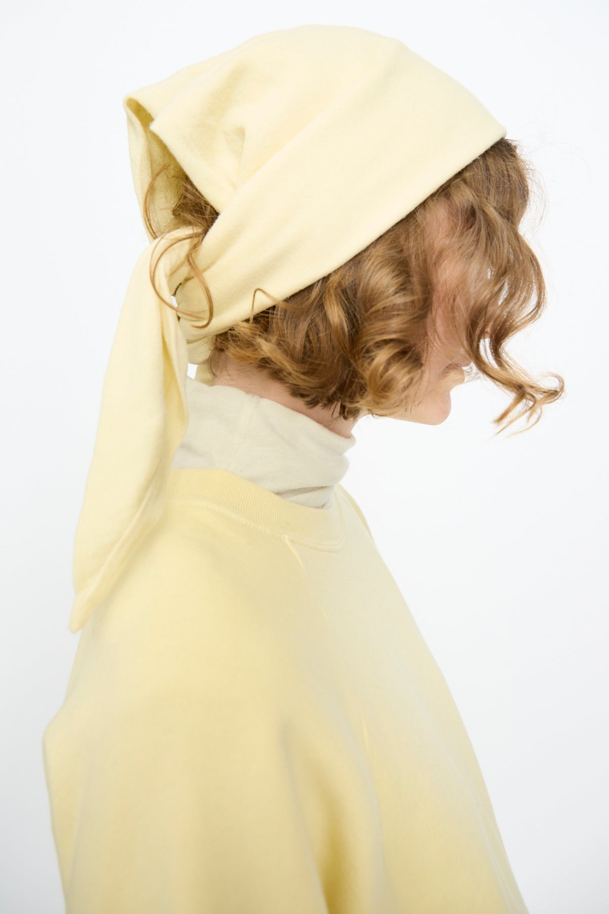 A person is shown in profile against a plain white background, wearing the Black Crane Triangle Scarf in Custard—a light yellow accessory crafted from a luxurious cotton cashmere blend for comfort and style—paired with a matching top.