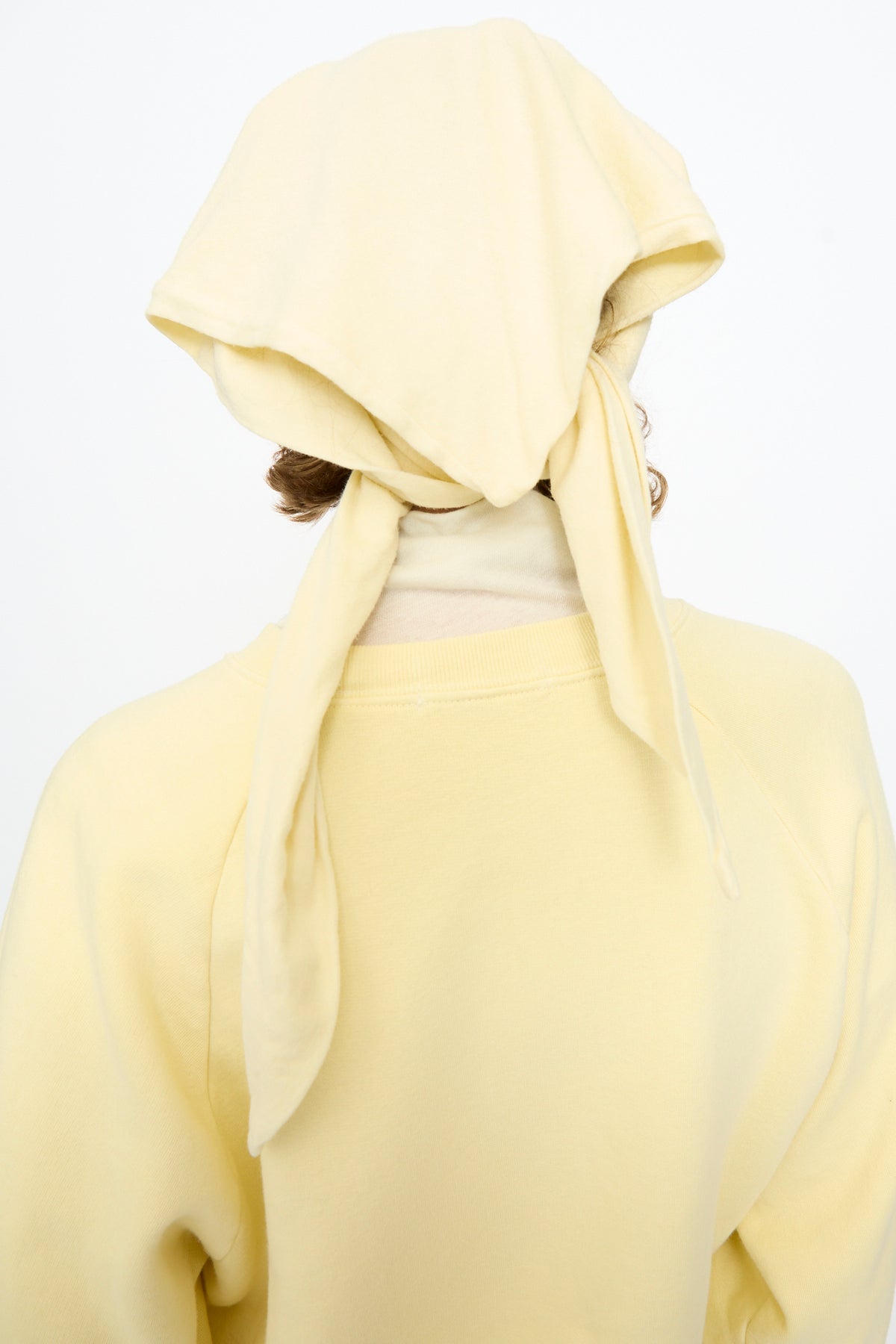 A person is seen from behind against a plain white background, wearing the Black Crane Triangle Scarf in Custard—a pale yellow piece crafted in the USA from a soft cotton-cashmere blend—and a matching top.