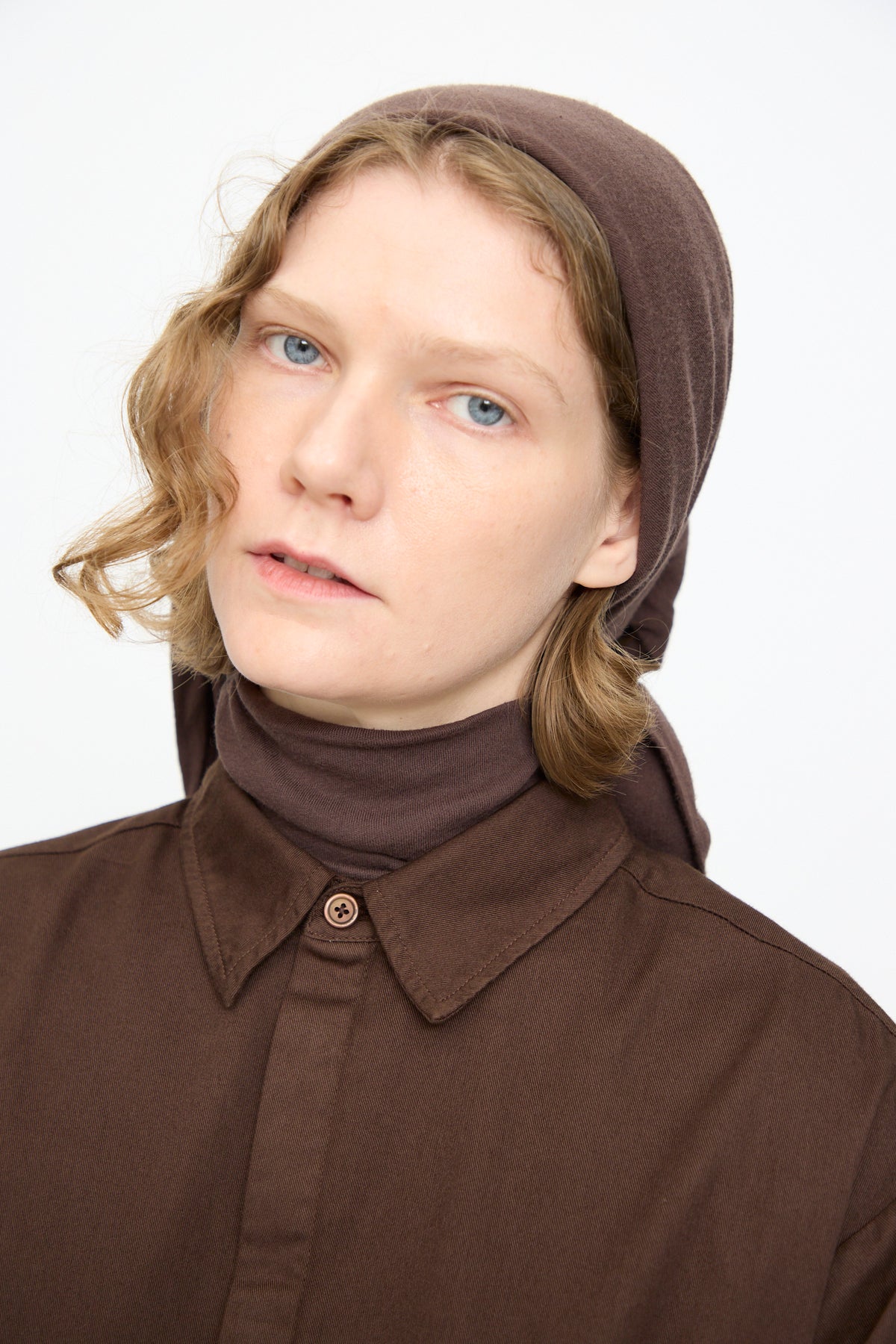A person wearing the Black Crane Triangle Scarf in Dark Plum and a brown button-up shirt looks at the camera with a neutral expression against a plain background. The scarf is crafted from soft cotton cashmere and made in the USA.