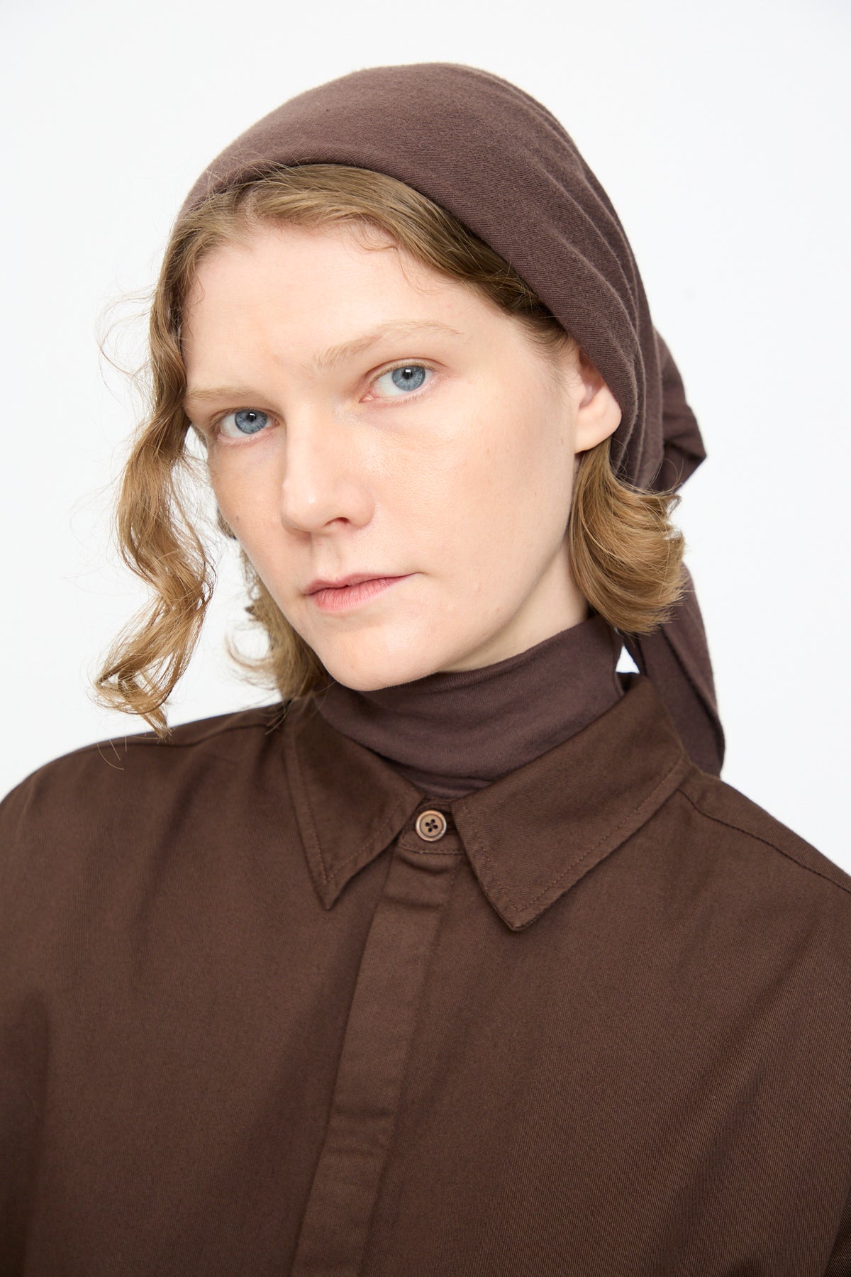 A person with light skin and wavy blonde hair wears the Black Crane Triangle Scarf in Dark Plum, made in the USA, along with a brown collared shirt, looking directly at the camera with a neutral expression against a plain background.