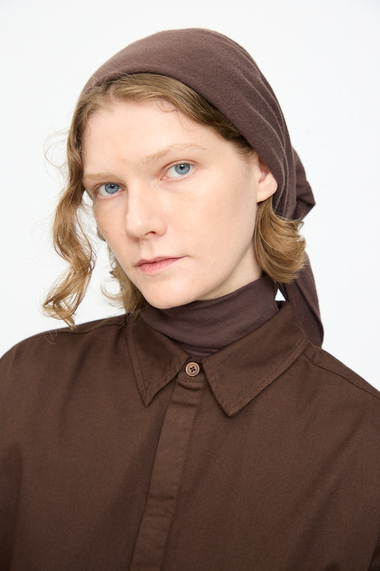A person with light skin and wavy blonde hair wears the Black Crane Triangle Scarf in Dark Plum, made in the USA, along with a brown collared shirt, looking directly at the camera with a neutral expression against a plain background.