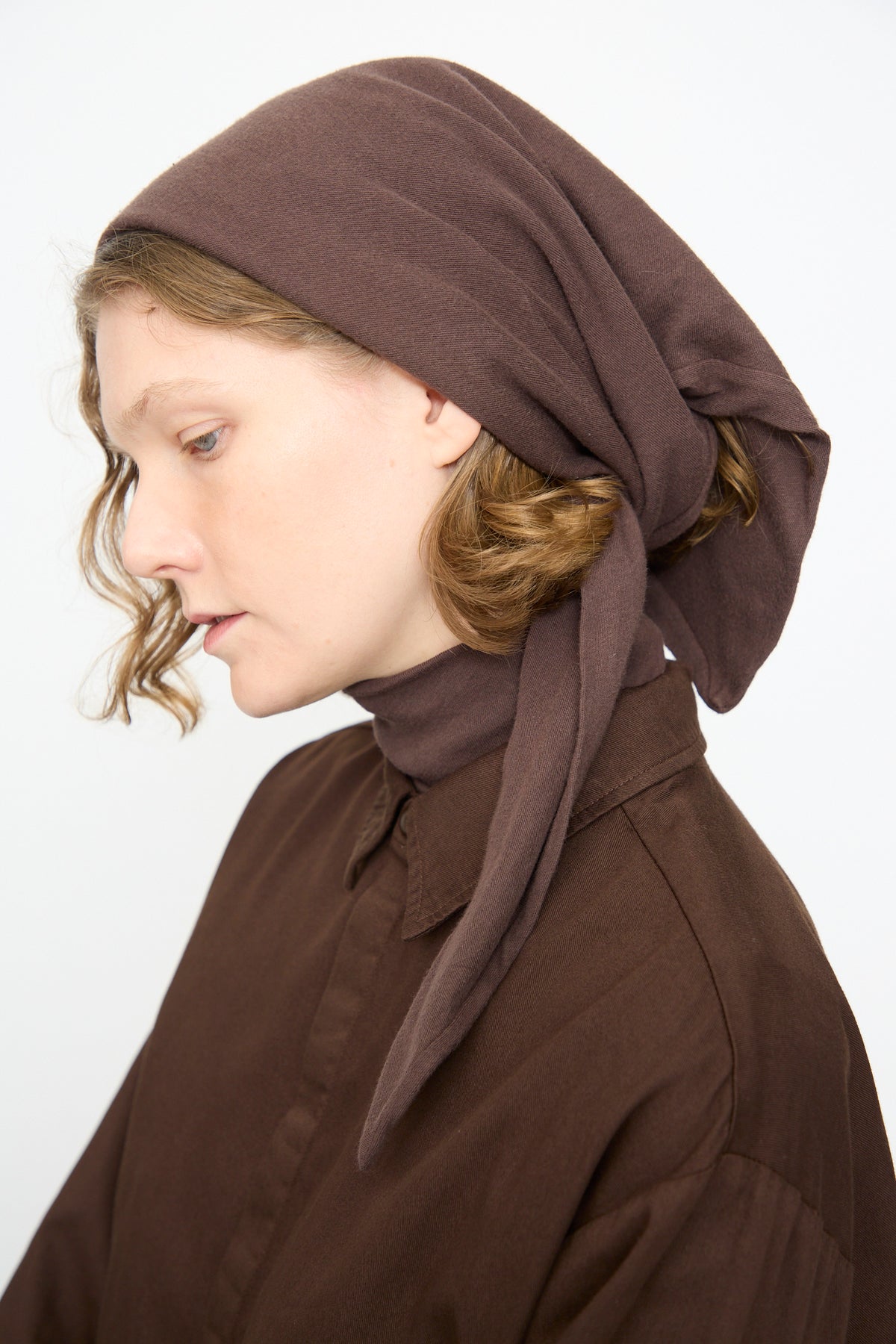 A woman in profile wears the Black Crane Triangle Scarf in Dark Plum styled with a brown collared shirt, looking down against a plain light background. The cotton cashmere scarf lends an elegant touch to her understated style.