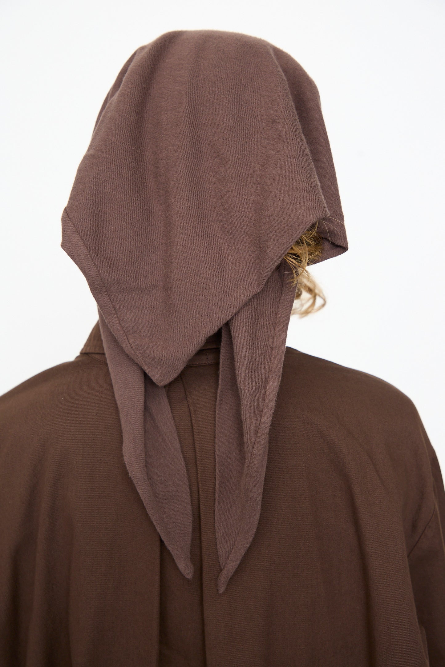 A person facing away, wearing the Black Crane Triangle Scarf in Dark Plum—crafted from luxurious cotton cashmere—and matching brown clothing; some curly hair showing beneath the headscarf; set against a plain white background.