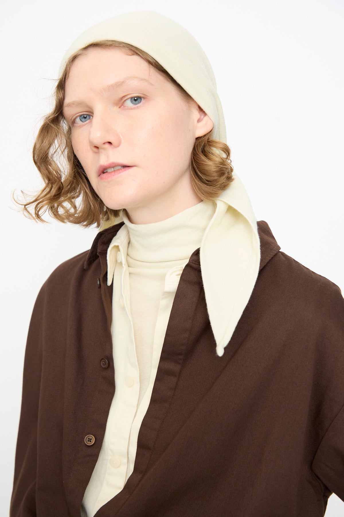 A person with short wavy hair wears the Black Crane Triangle Scarf in Natural, paired with a cream turtleneck and a brown button-up shirt, looking at the camera against a plain white background.