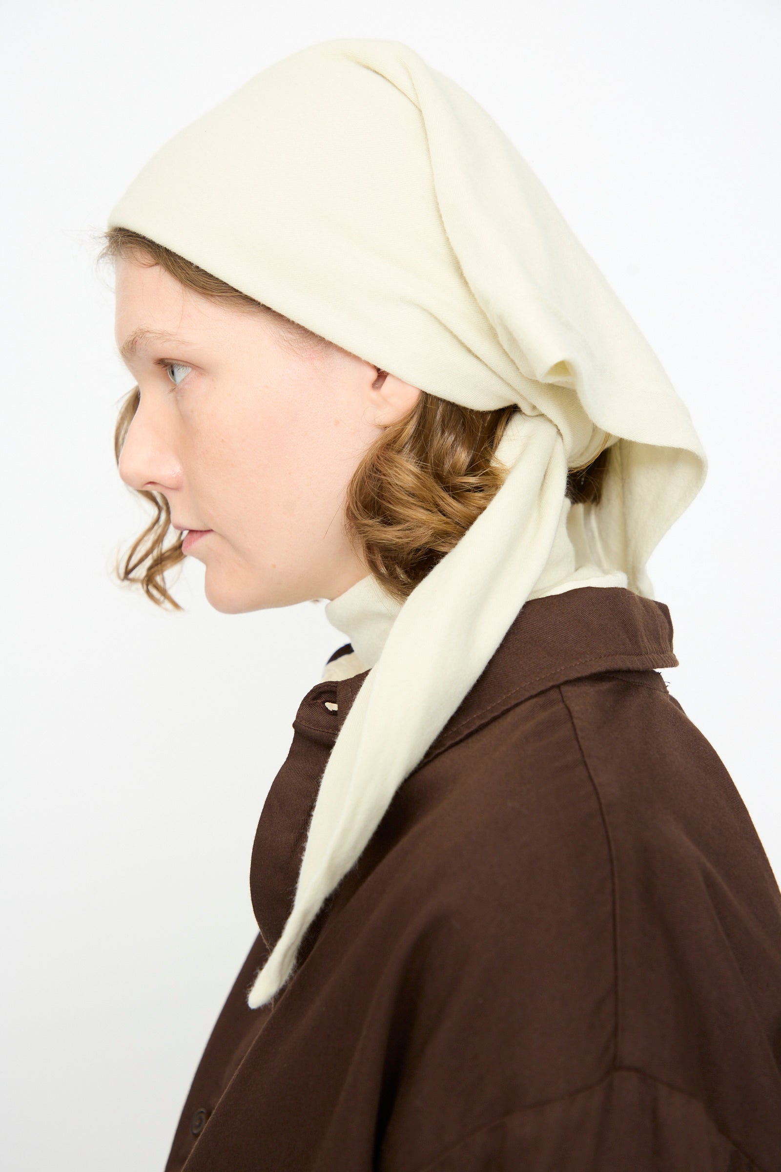 A woman with light skin and curly hair is shown in profile against a plain white background, wearing a brown coat and the Black Crane Triangle Scarf in Natural—a luxurious cotton cashmere blend scarf made in the USA.